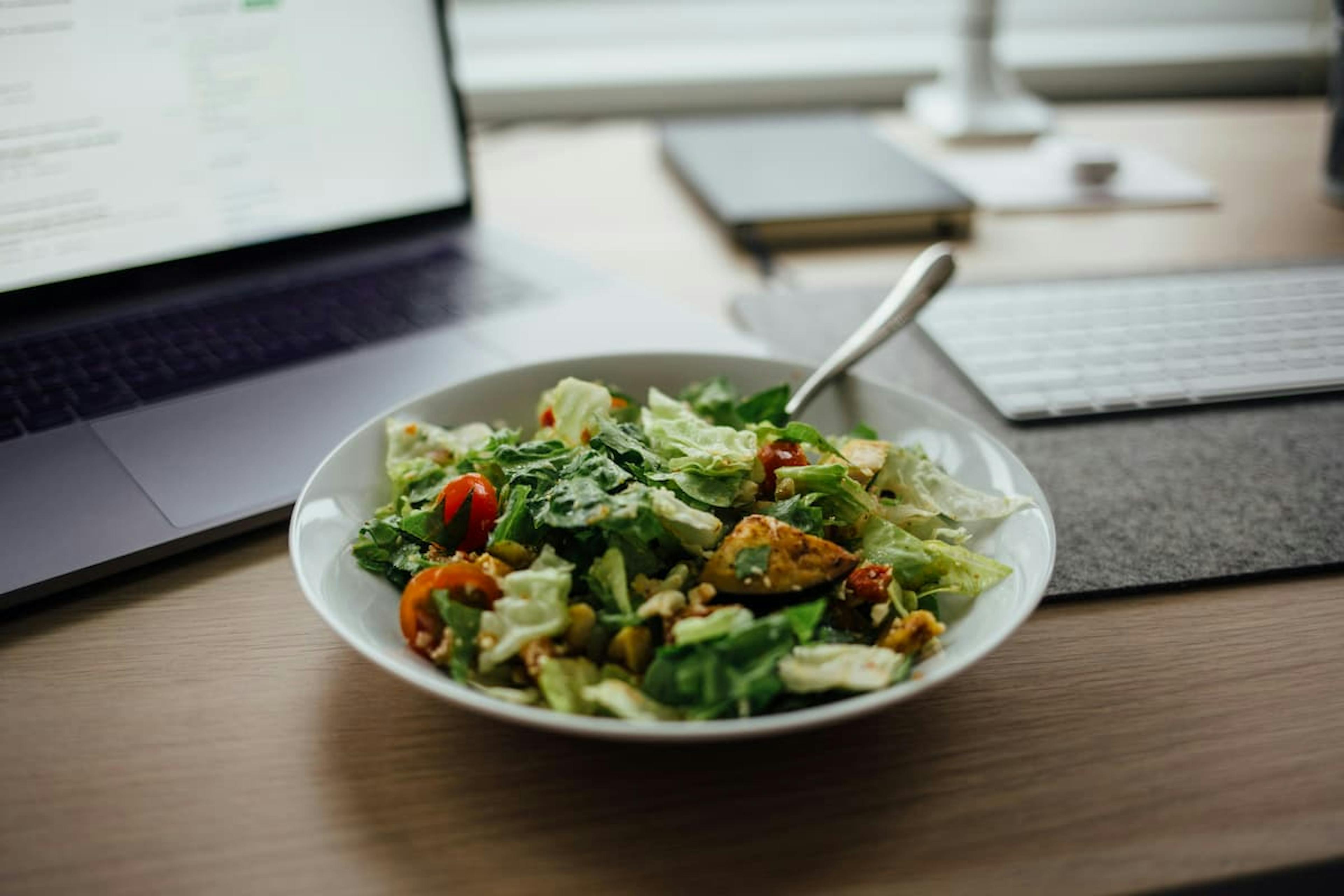 A bowl of fresh salad with lettuce, cherry tomatoes, grilled chicken, and mixed greens on an office desk next to a laptop and keyboard.