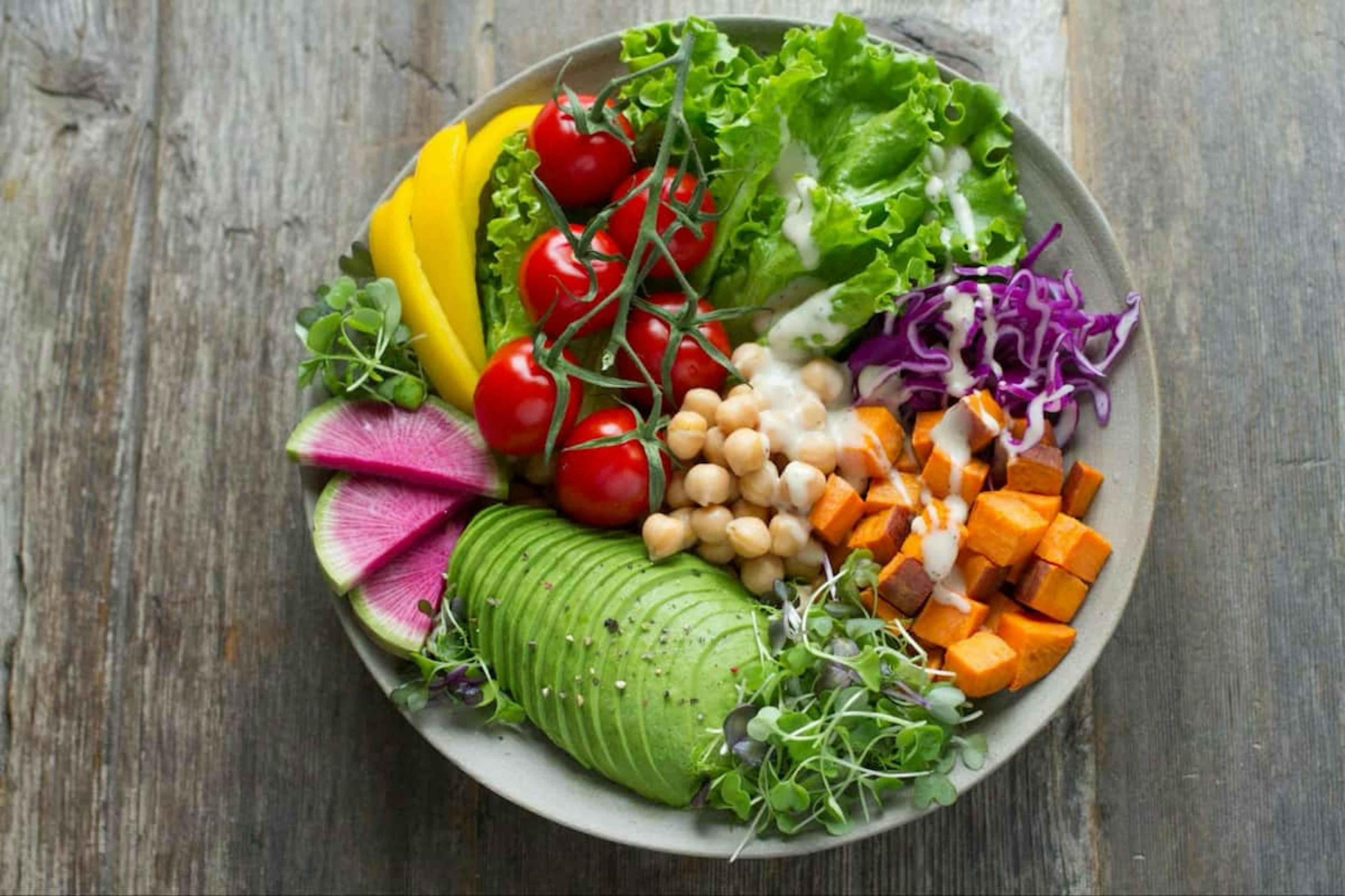 A colorful salad bowl with sliced avocado, cherry tomatoes on the vine, yellow bell pepper, lettuce, chickpeas, cubed sweet potatoes, red cabbage, radish slices, and microgreens, drizzled with a light dressing.