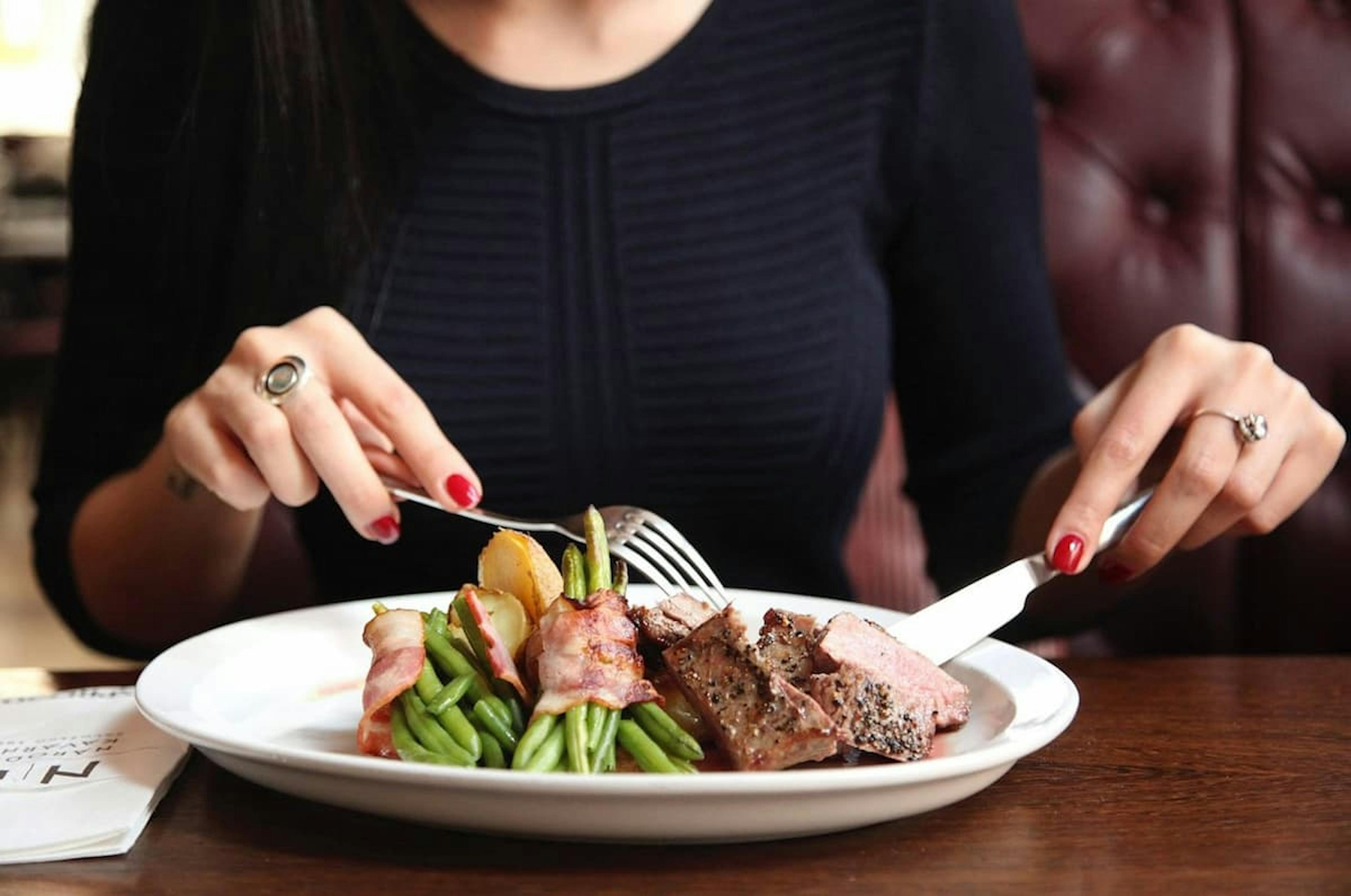 Person enjoying a plate of healthy food with meat, green beans wrapped in bacon, and potatoes at a restaurant.