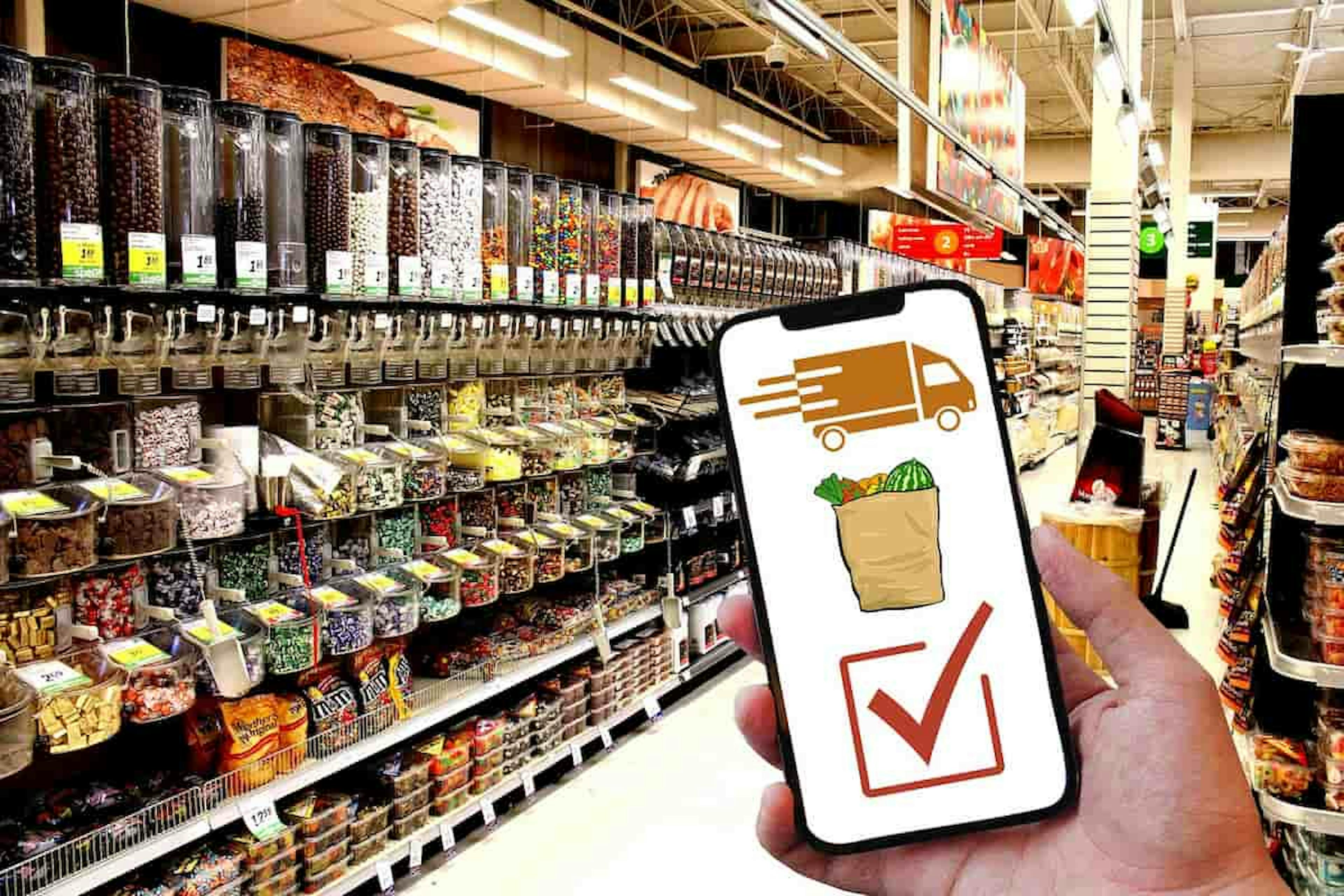 Hand holding a smartphone with grocery delivery app open in a supermarket aisle filled with bulk food containers and snack options.