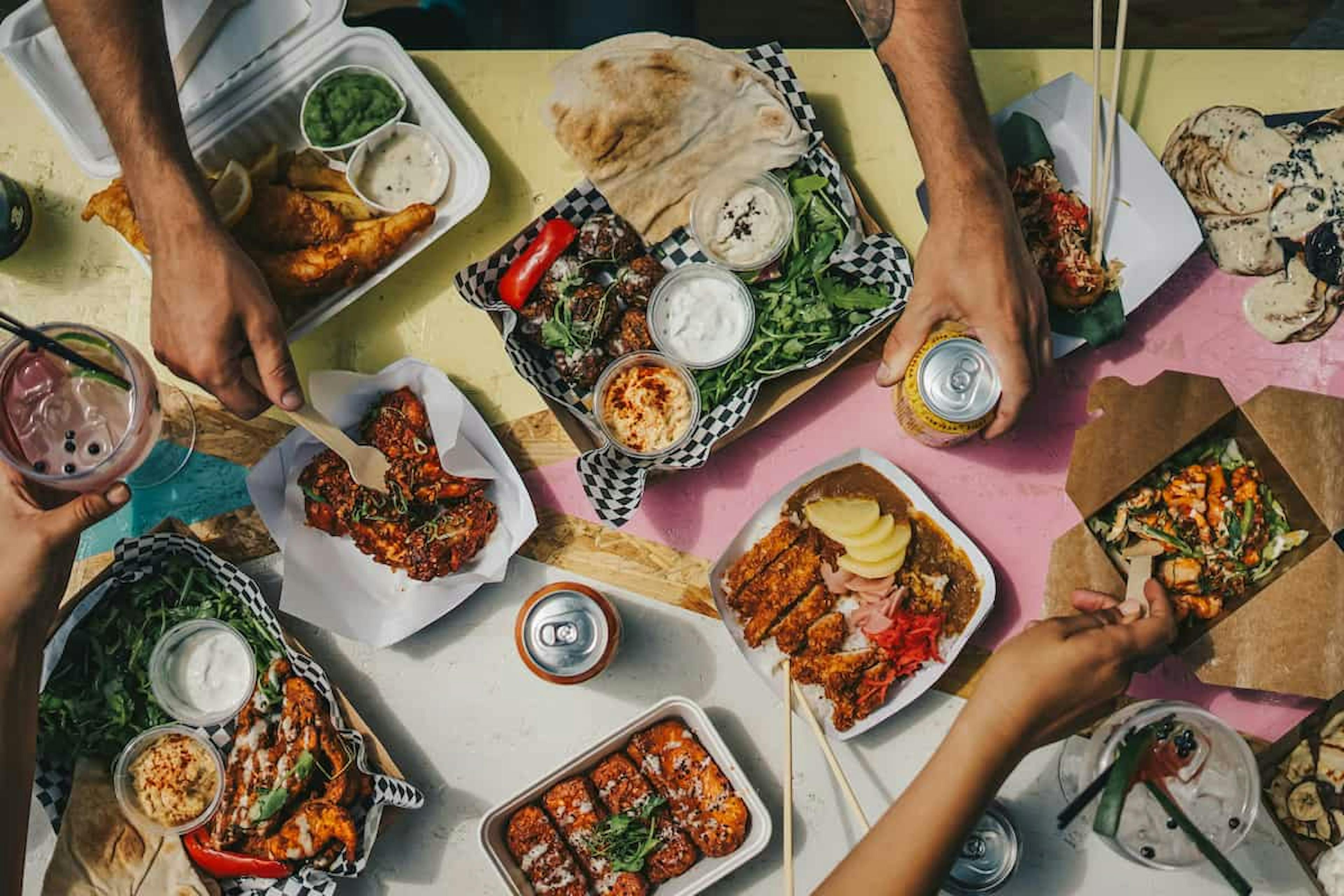 A vibrant assortment of dishes on a shared table, including fried chicken, dips, grilled vegetables, pita bread, and a variety of international flavors, with hands reaching in to share the meal.