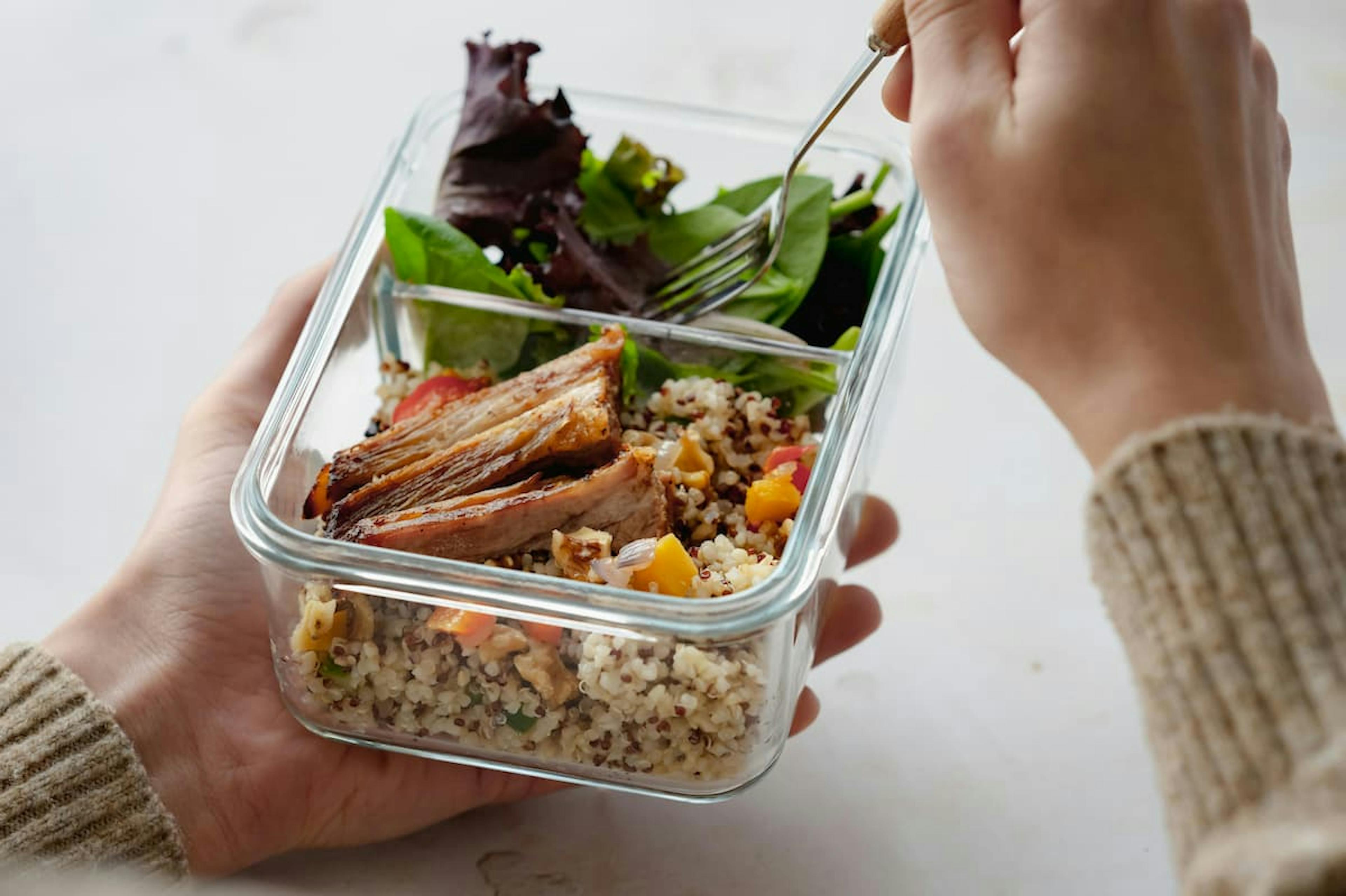 Close-up of a glass container with a healthy meal, including mixed quinoa, grilled meat, and fresh greens. A person is holding the container and using a fork to eat.