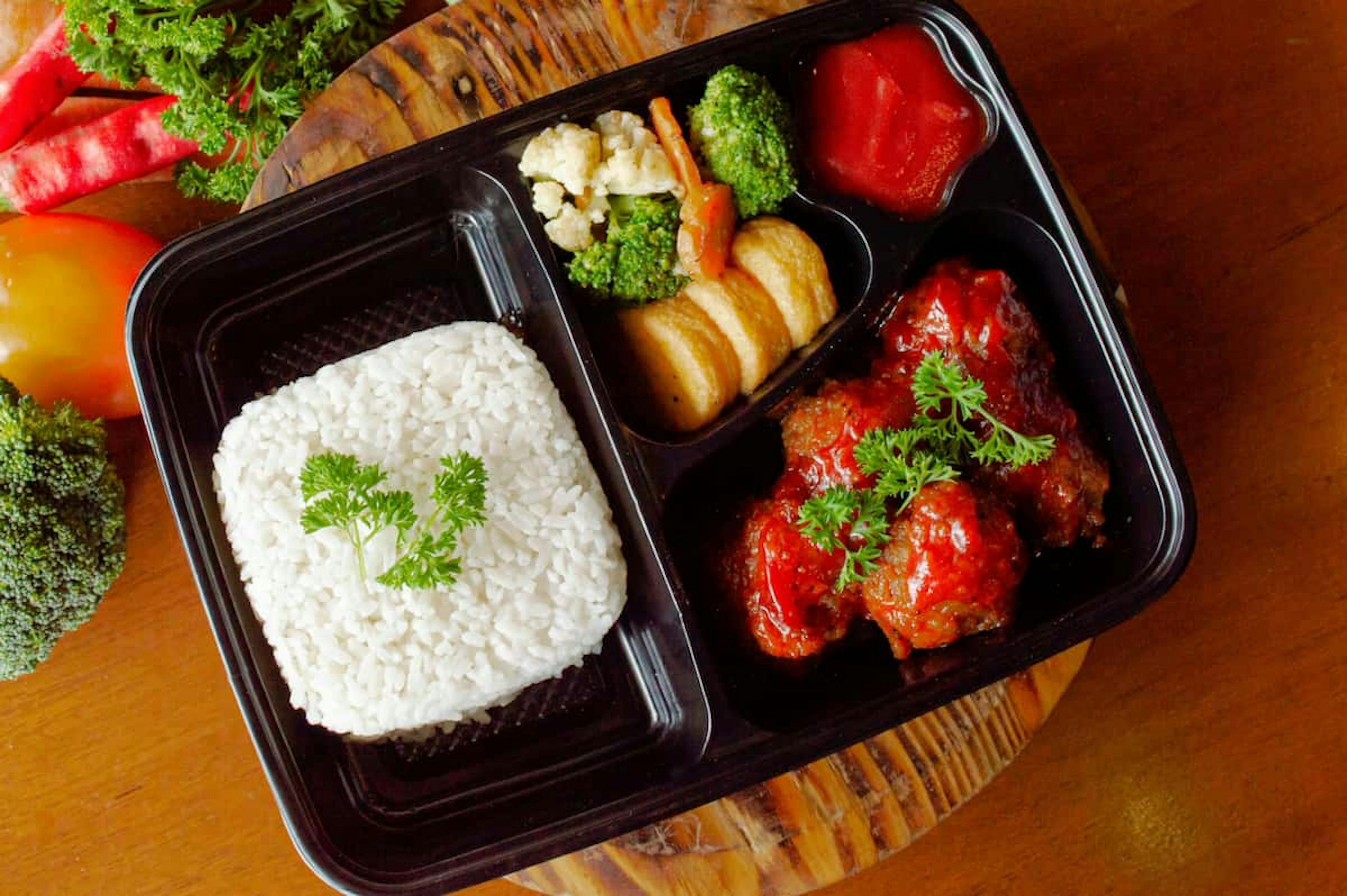 A black meal tray with rice, vegetable medley, fried potatoes, and a meat dish garnished with parsley, placed on a wooden table with fresh vegetables around.