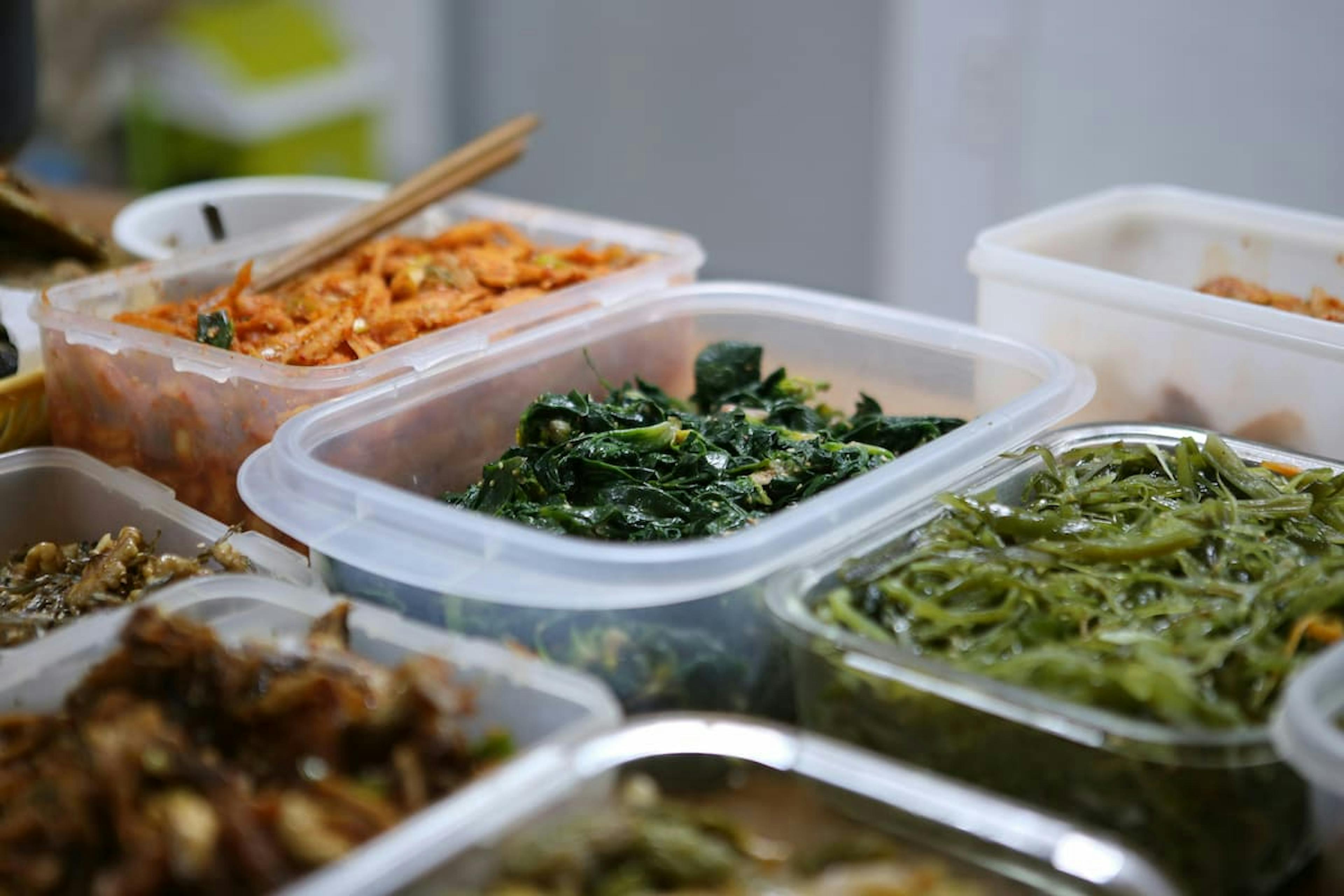 Various fresh vegetables in plastic containers prepared for meal planning or storage, including seasoned greens, shredded carrots, and sautéed greens.