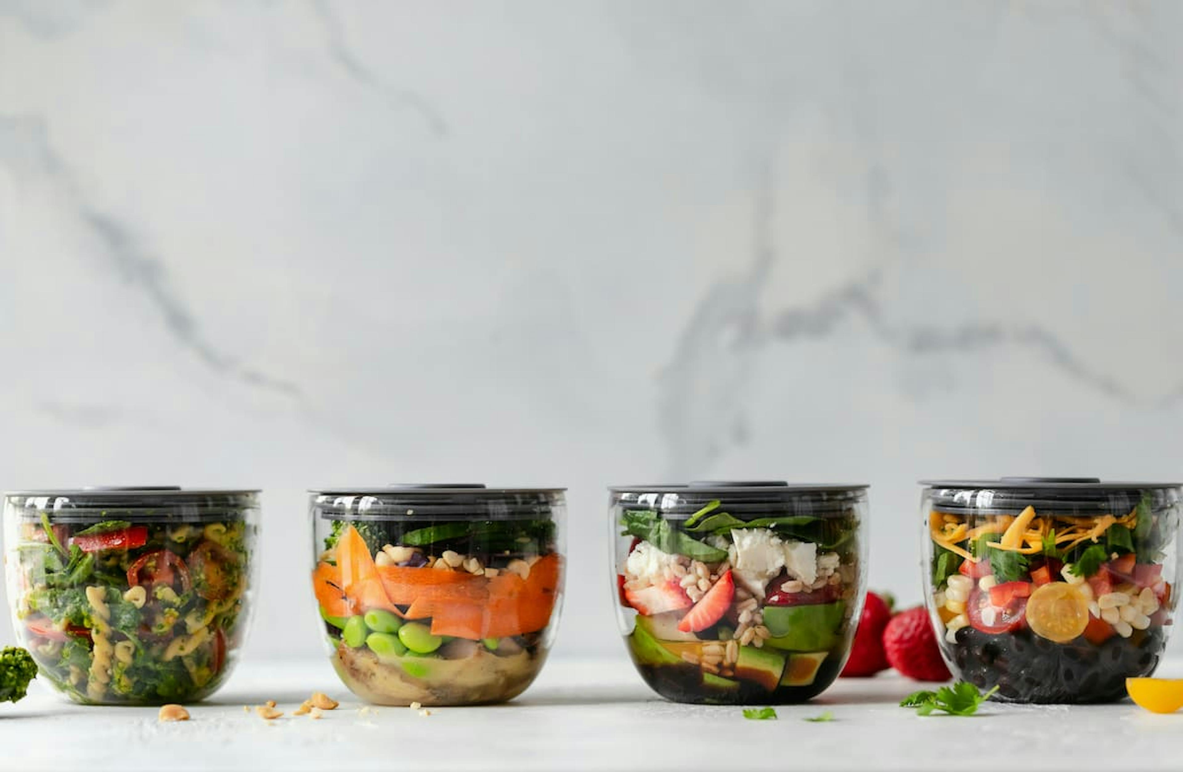 Four glass containers with colorful, layered salad ingredients, including greens, grains, vegetables, and fruits, prepared for meal planning.