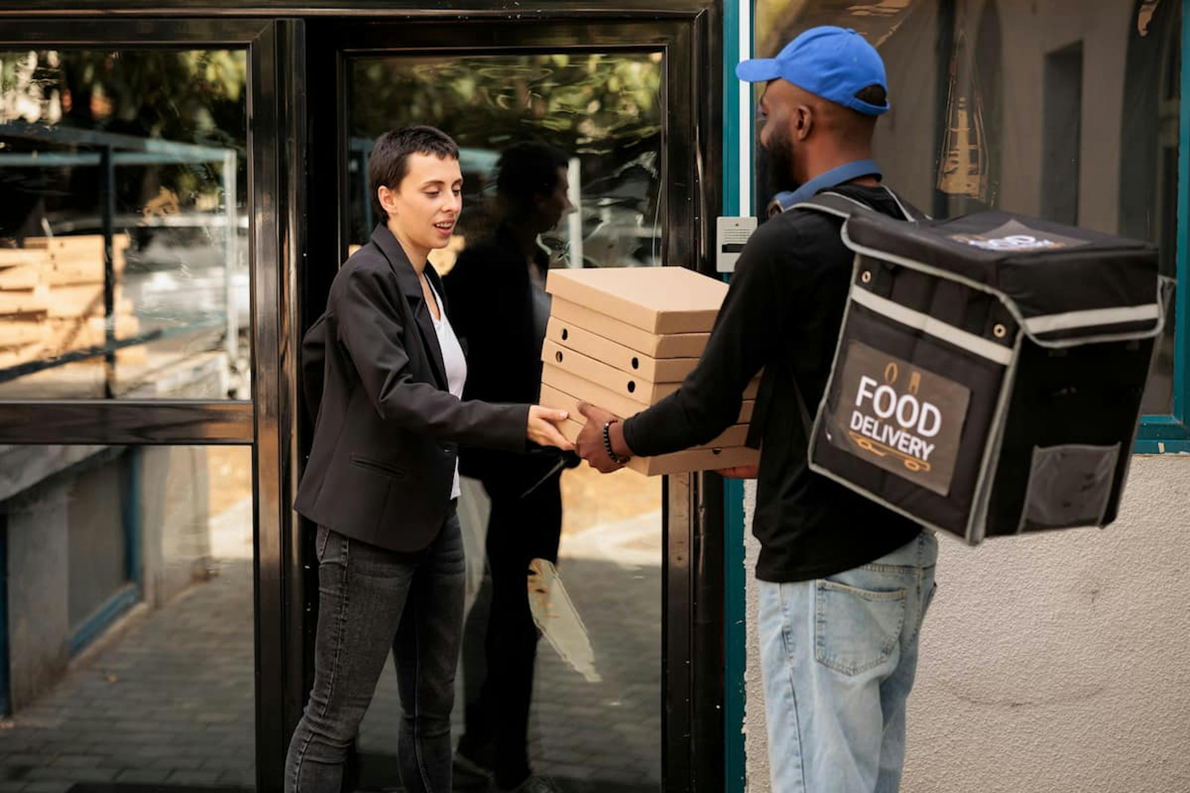 A food delivery person hands a stack of pizza boxes to a businesswoman outside a modern office building, representing meal delivery service.