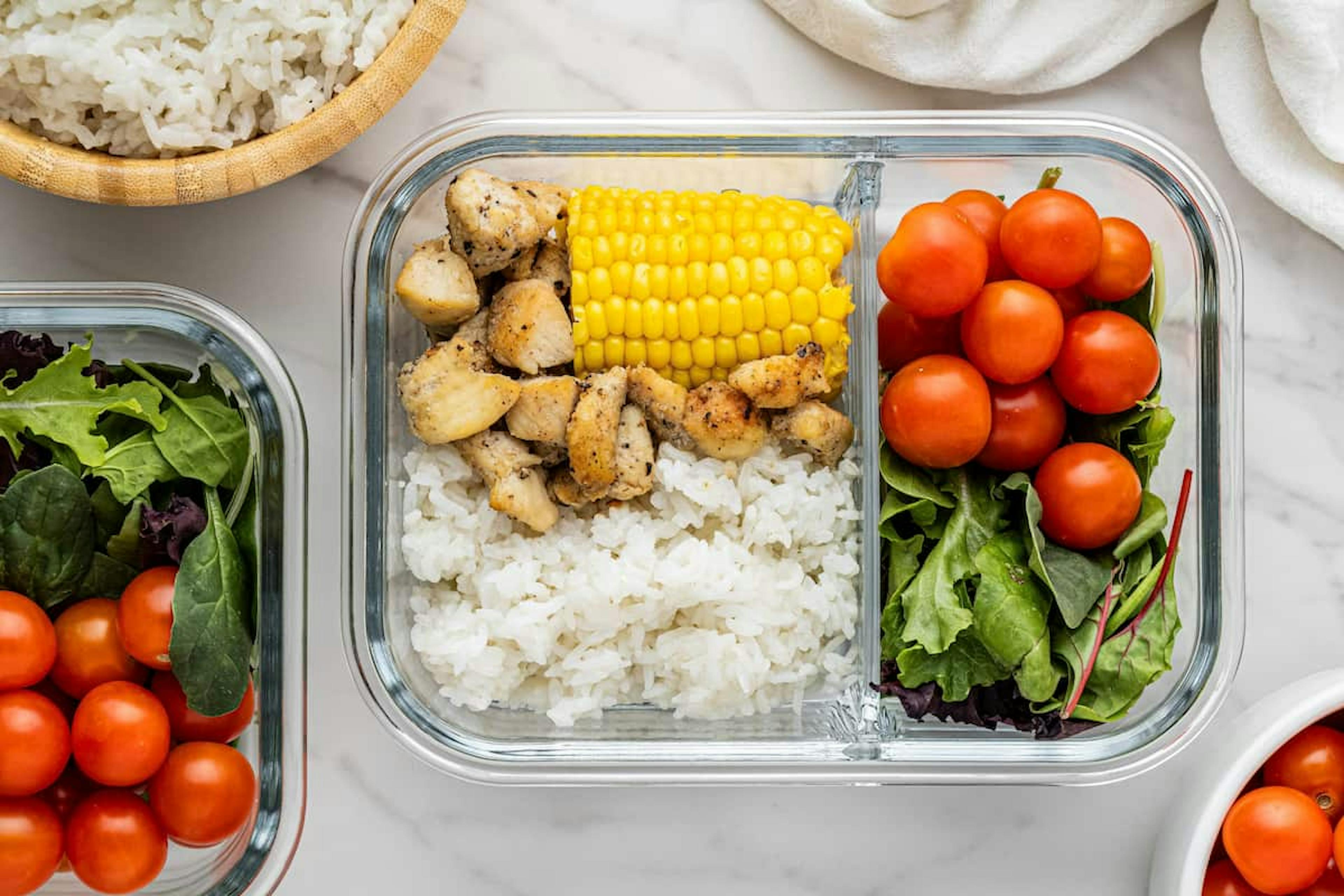 A glass meal prep container with grilled chicken, rice, corn on the cob, cherry tomatoes, and mixed greens, representing a healthy lunch option.