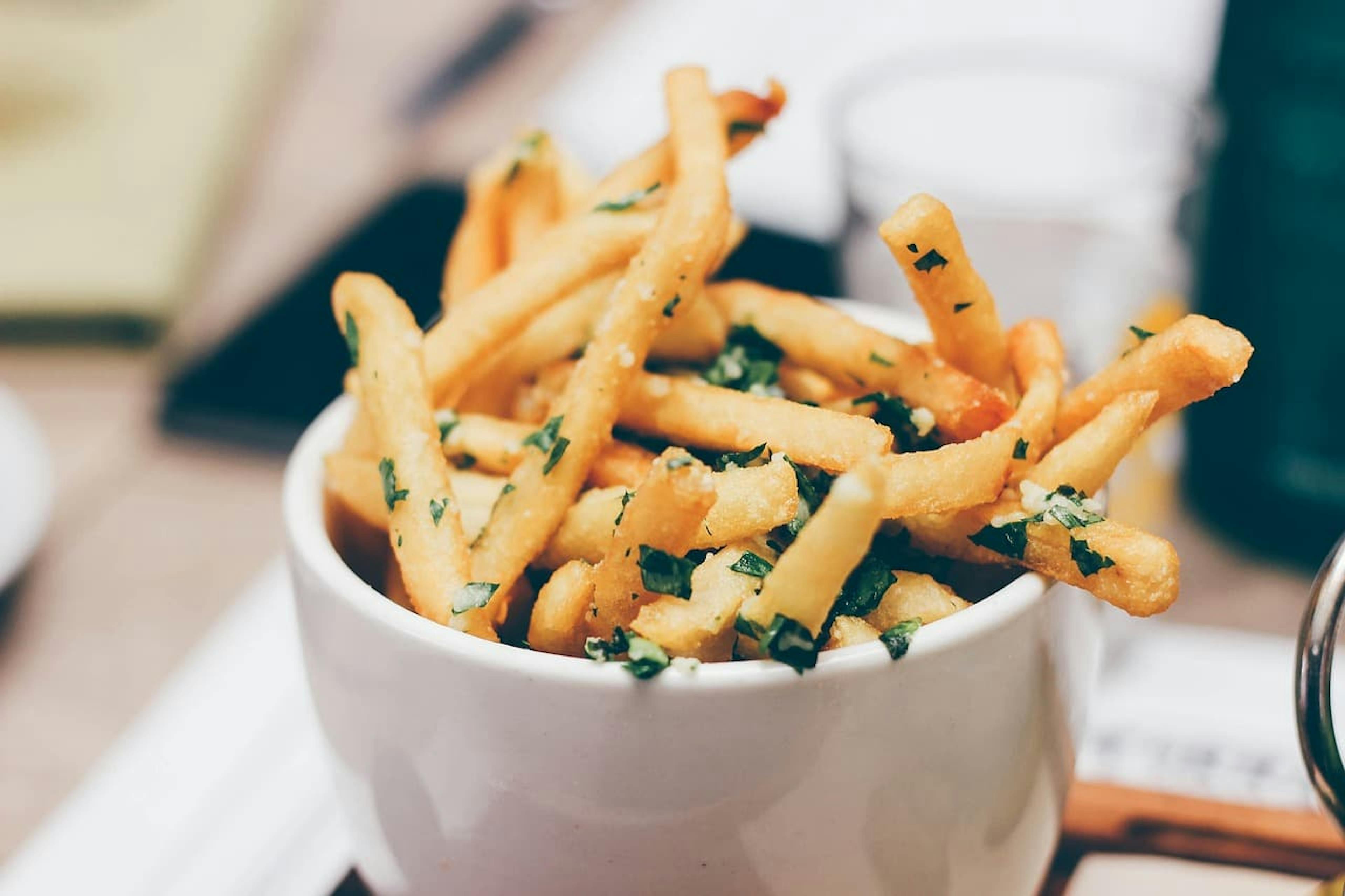 White bowl filled with golden French fries garnished with chopped parsley, placed on a blurred dining table background.