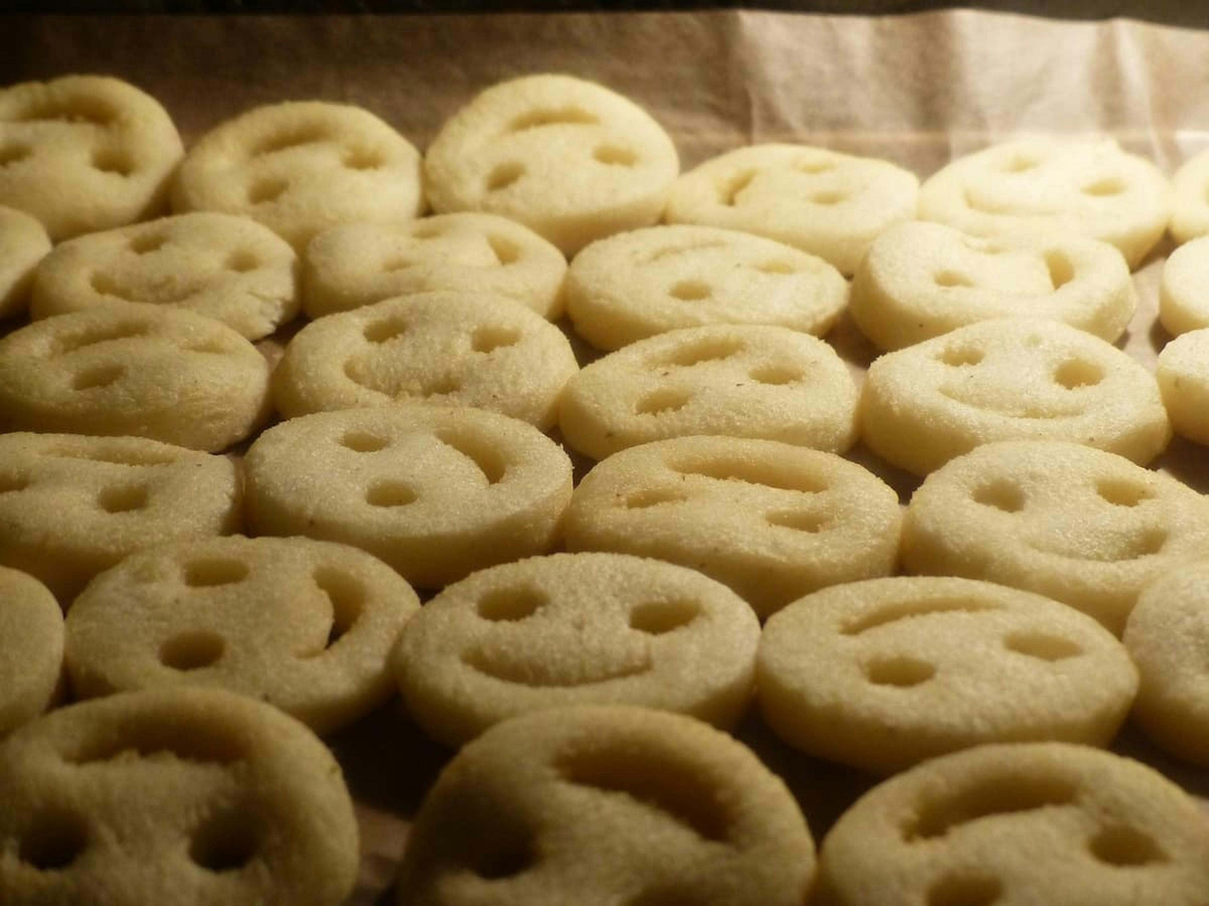 "Tray of frozen potato smiley faces with expressive cut-out eyes and mouths, arranged in rows on parchment paper.