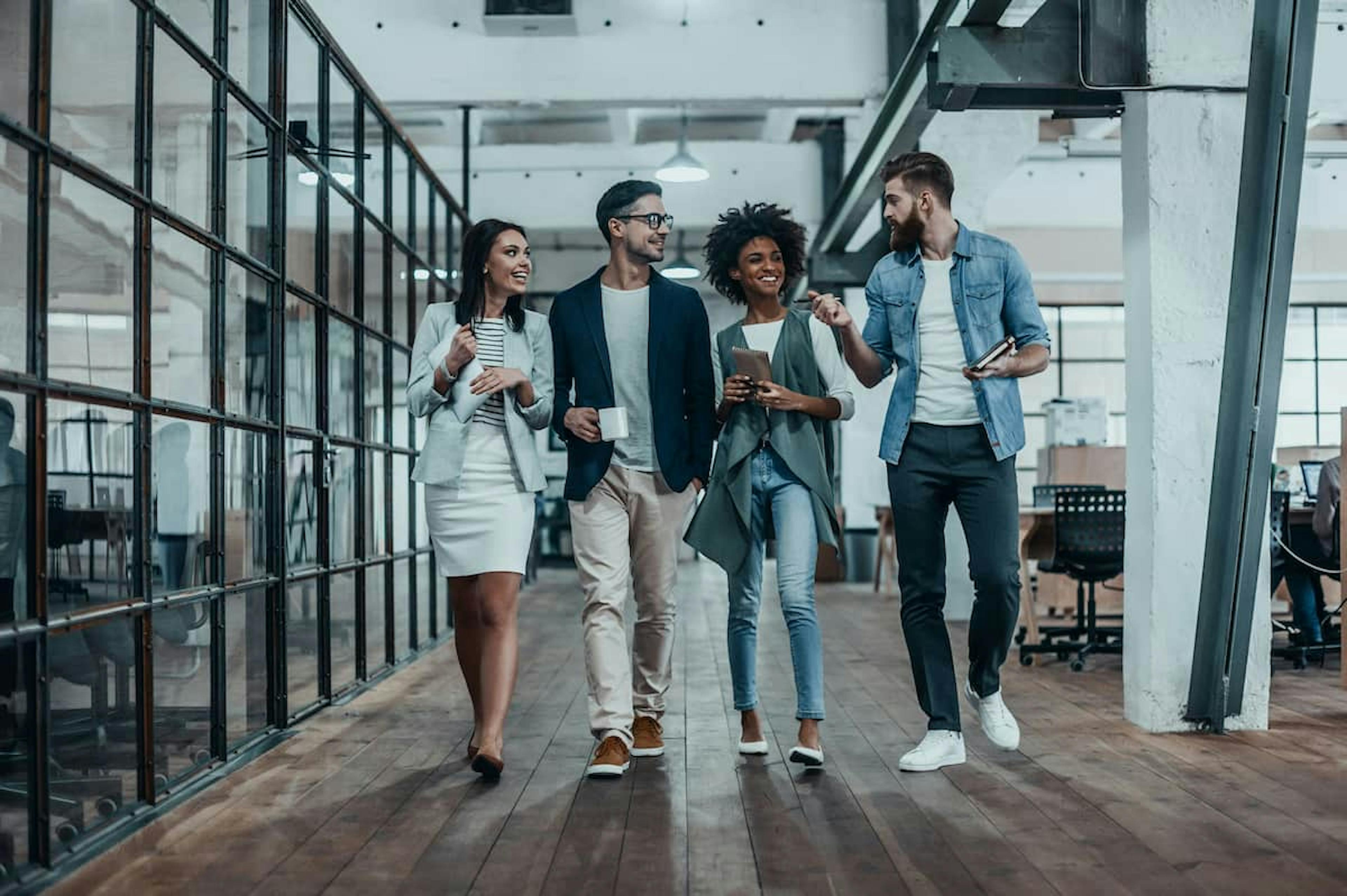 Four colleagues walking and talking in a modern office space, smiling and holding coffee cups and notebooks, fostering teamwork and engagement.