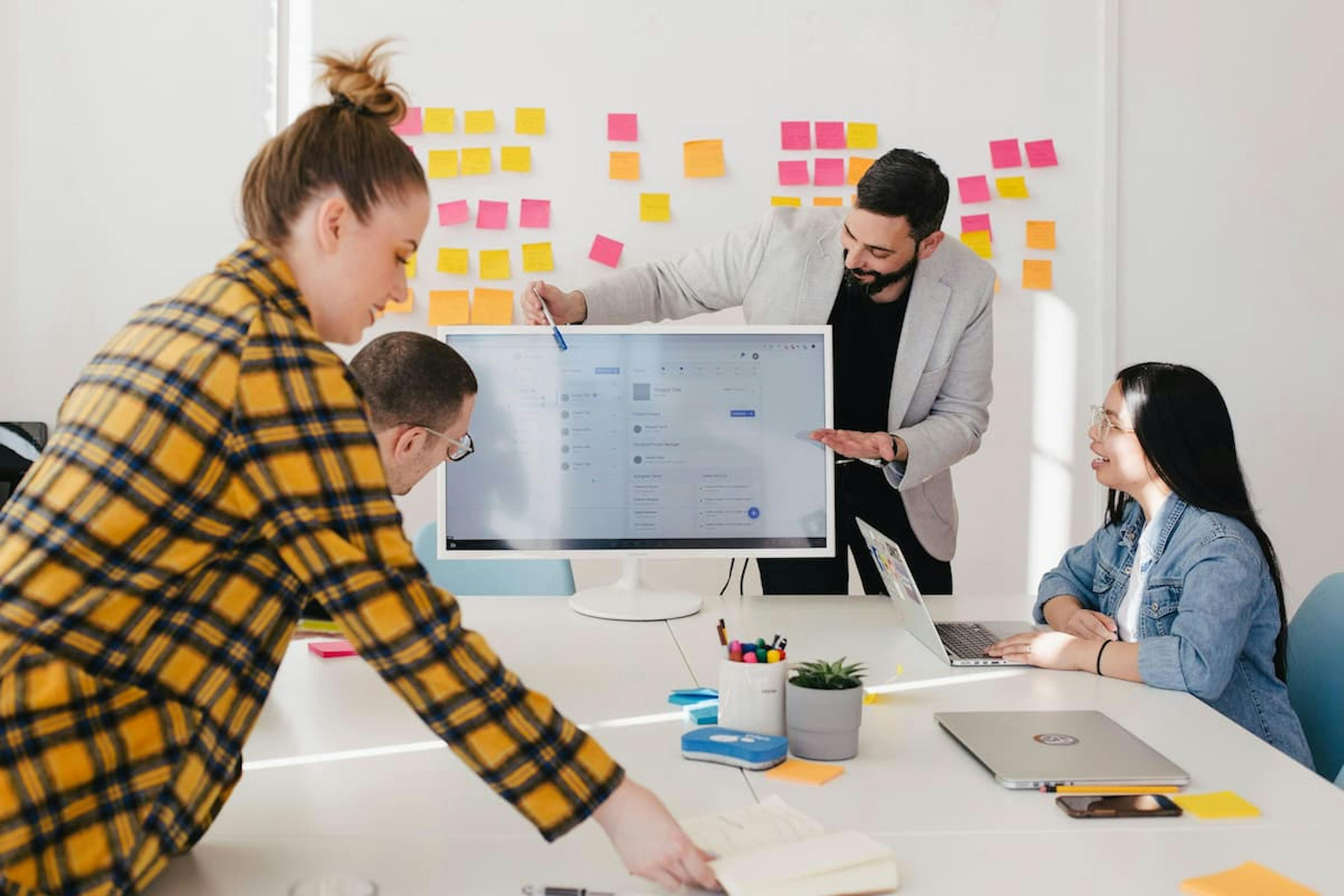 Team collaborating in a bright office, with a man presenting on a computer screen and colorful sticky notes on the wall.