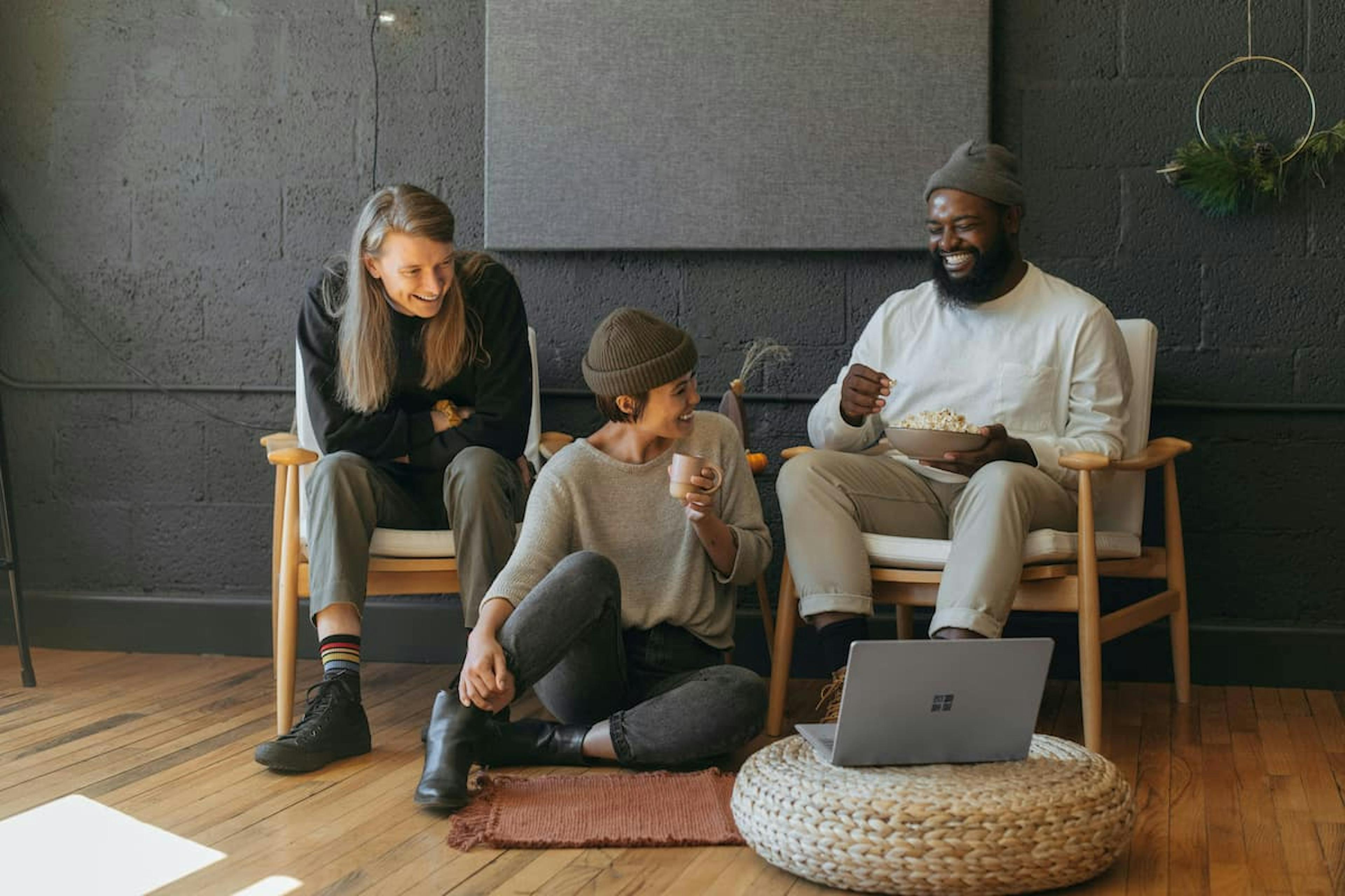 Three people sitting and smiling in a cozy indoor setting. Two are seated on chairs, and one is sitting on the floor holding a cup, while another holds a bowl of popcorn. A laptop rests on a pouf in front of them, creating a relaxed and casual atmosphere.