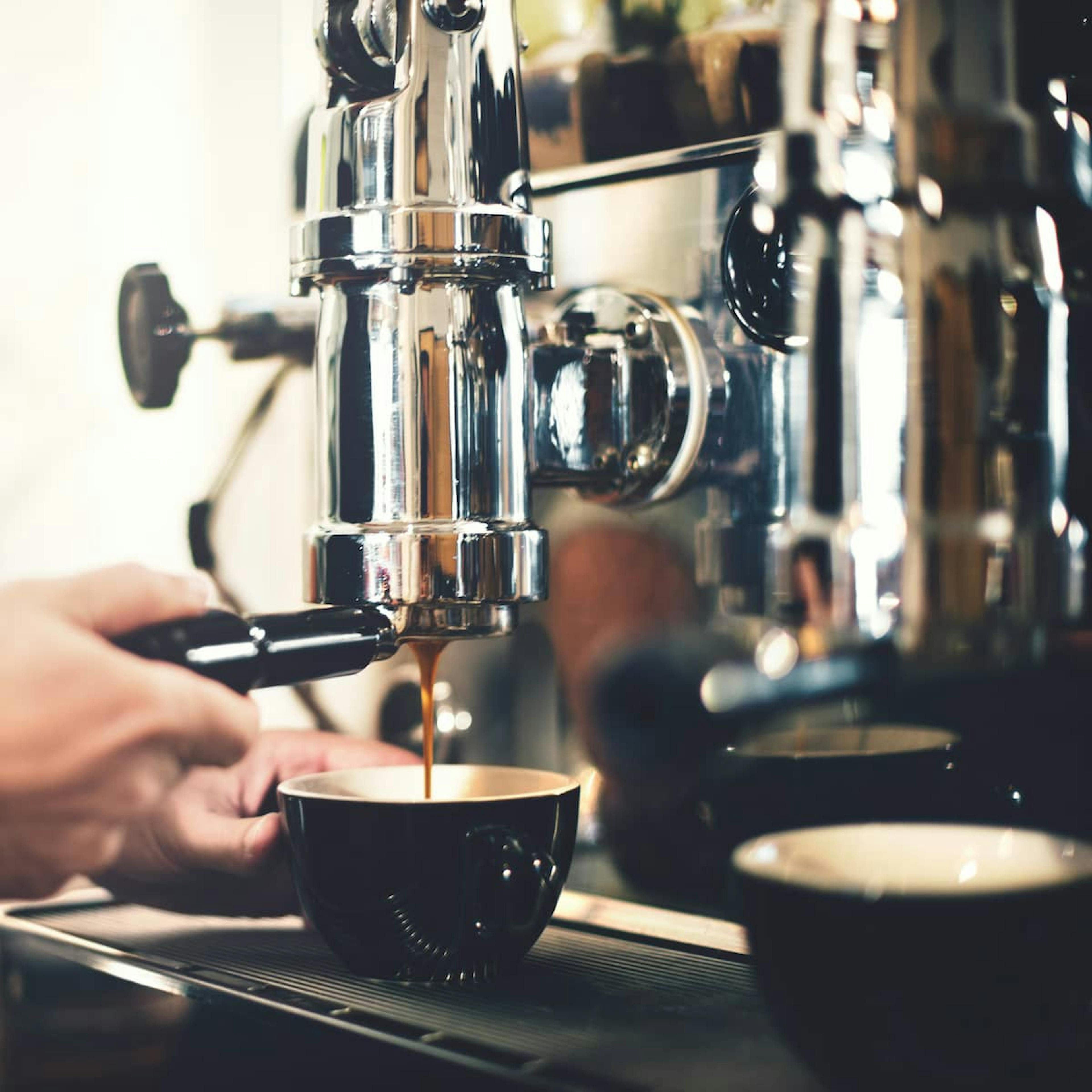 Close-up of a professional espresso machine brewing coffee into a black ceramic cup. A hand is visible holding the portafilter, with a shiny metallic surface reflecting the surroundings.