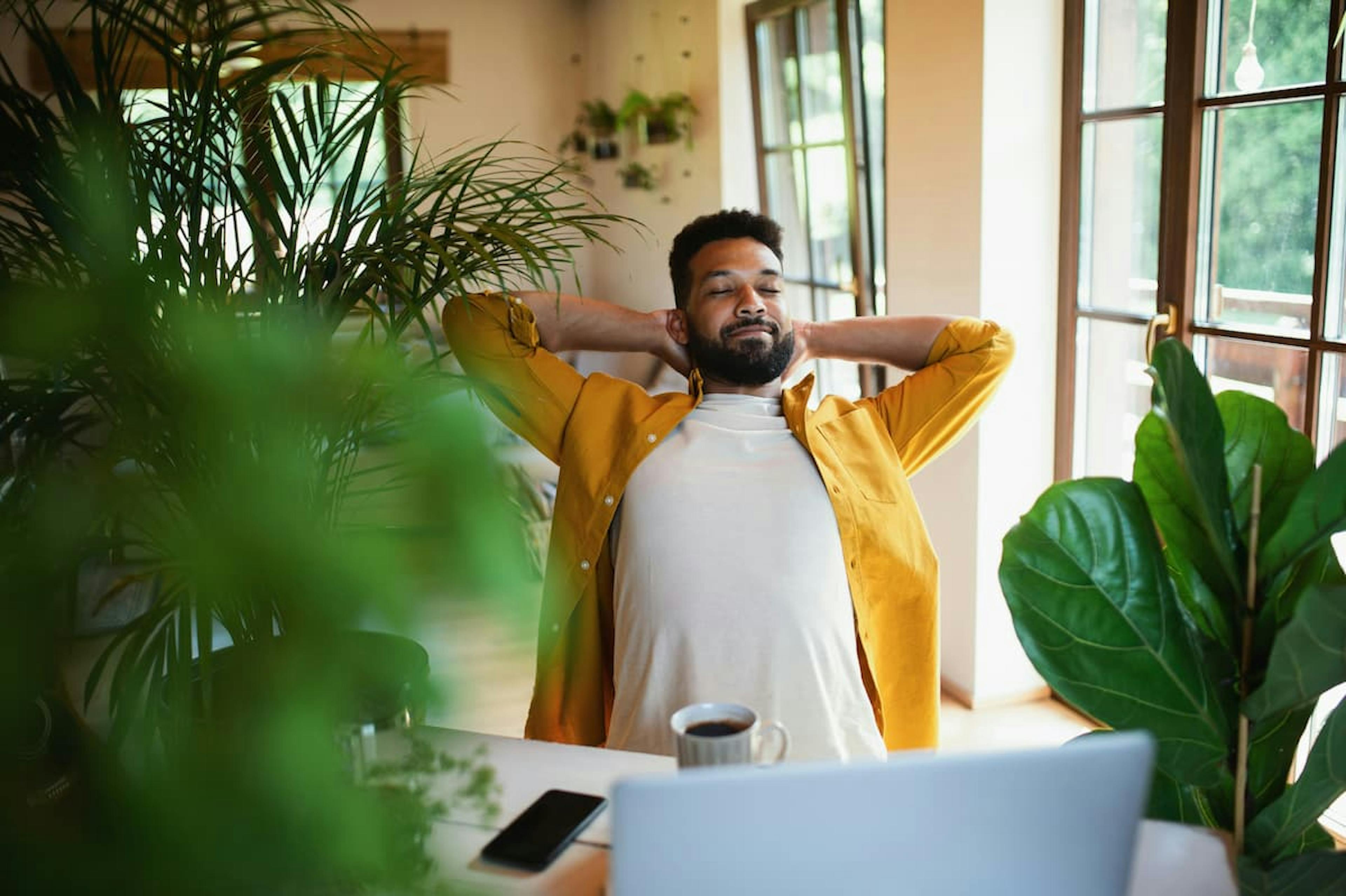 Man in a bright yellow shirt sitting at a desk, leaning back with hands behind his head and eyes closed, enjoying a moment of relaxation. A coffee mug and laptop are visible on the desk, surrounded by lush green indoor plants.