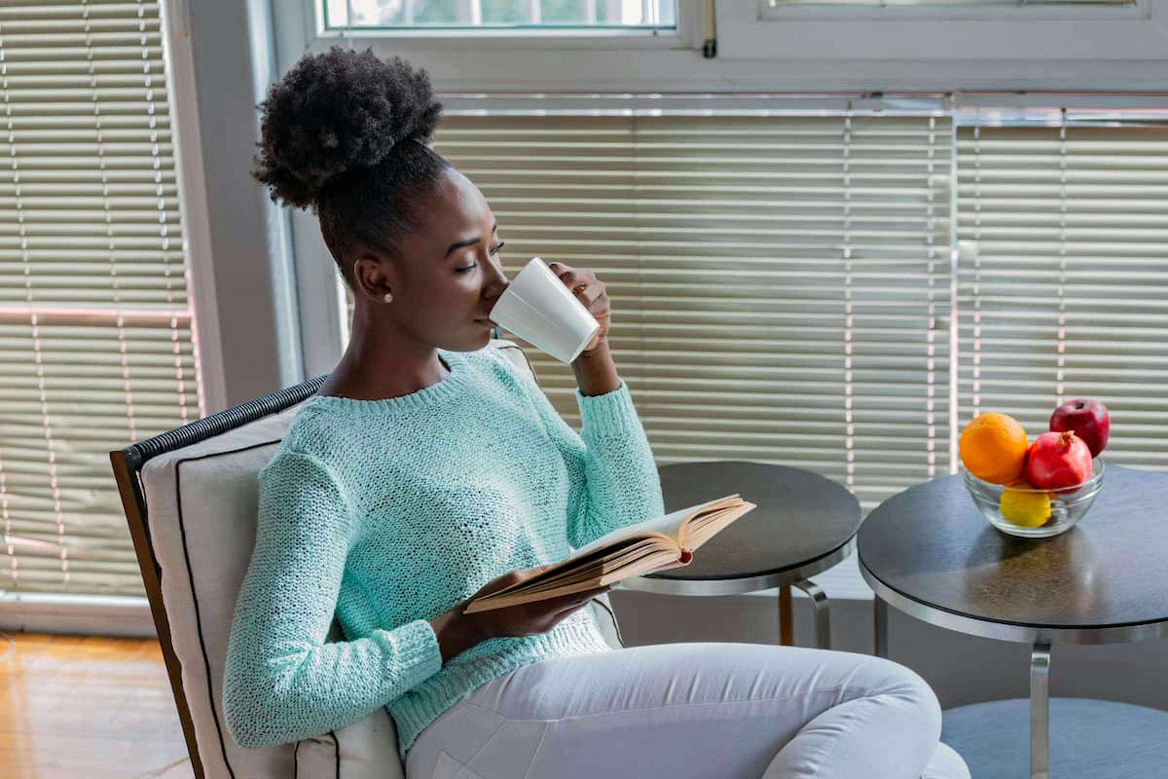 A woman sits in a cozy chair near a window with blinds, wearing a light blue sweater and white pants, holding a book and sipping from a white mug. A bowl of fresh fruit, including oranges and apples, is on a side table nearby.