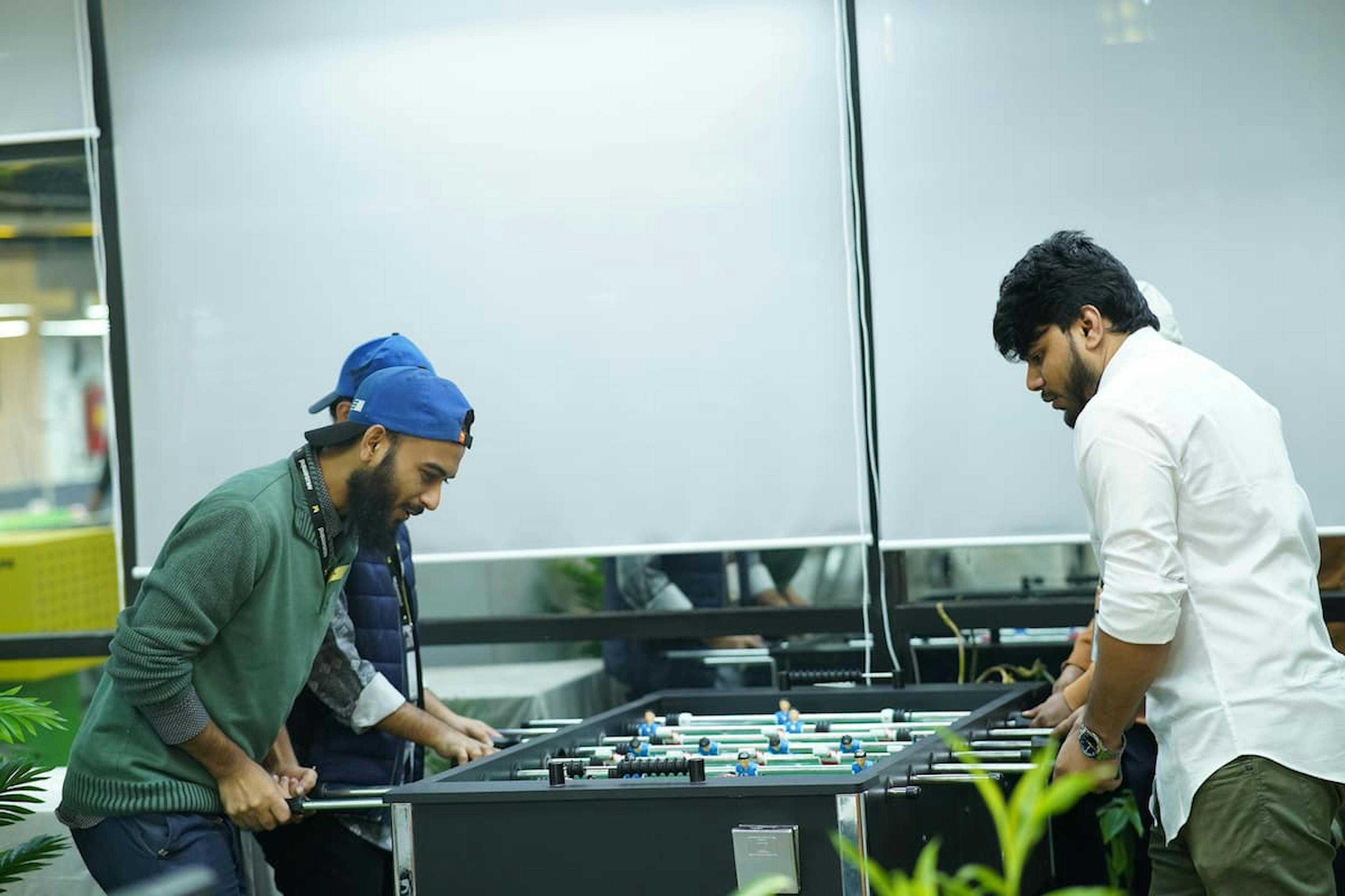 Two men playing a game of foosball in a modern indoor setting, one wearing a green sweater and blue cap and the other in a white shirt. The room features large windows with blinds and is decorated with greenery.