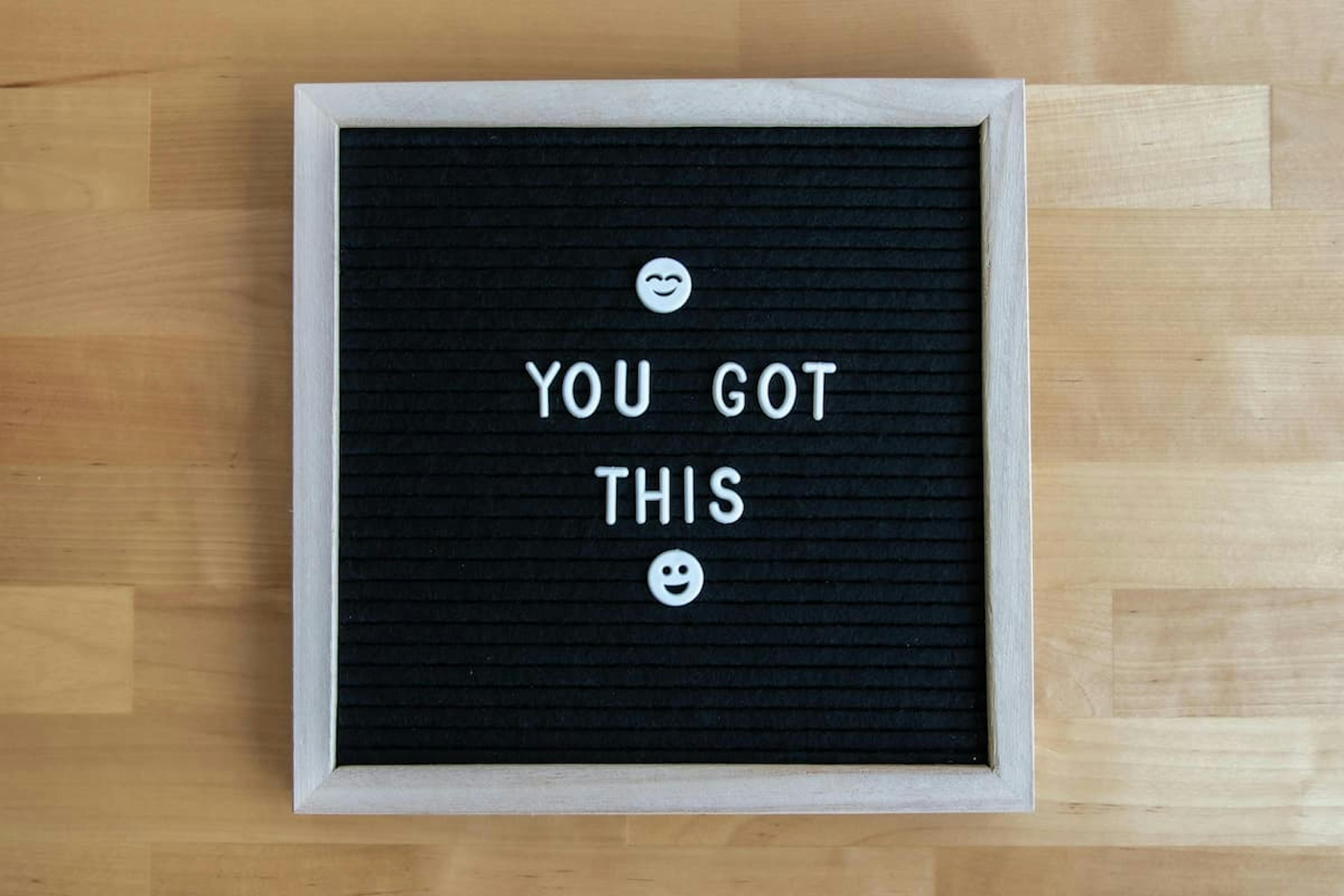 A motivational black letter board with a light wooden frame displaying the message "YOU GOT THIS" in white letters, accompanied by two smiley face emojis, placed on a light wooden surface.