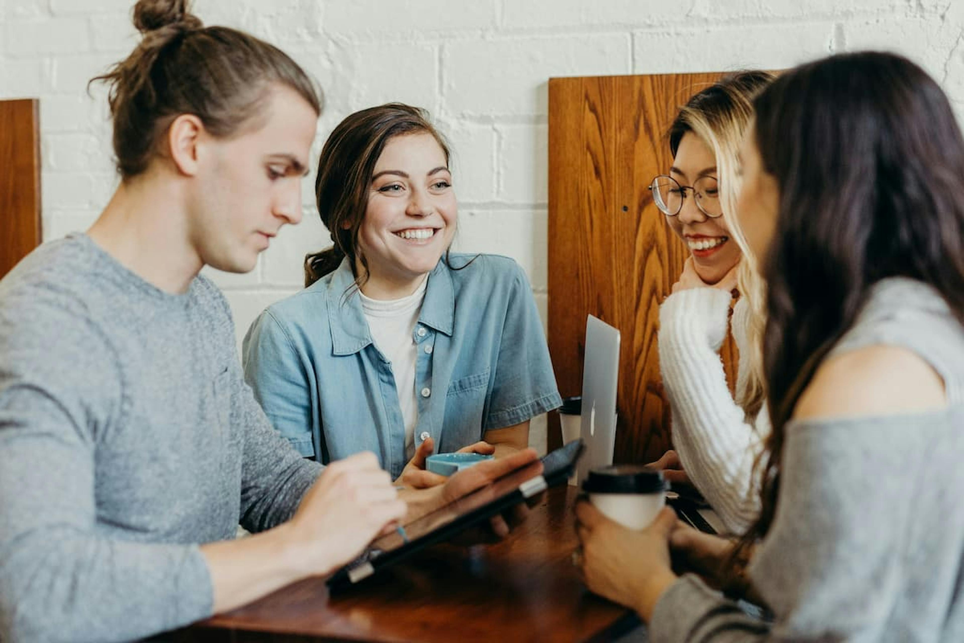 A group of four young adults sitting around a wooden table in a casual setting, engaged in a friendly conversation. One person is holding a tablet, another has a coffee cup, and a laptop is visible on the table. They are smiling and appear to be enjoying their interaction.