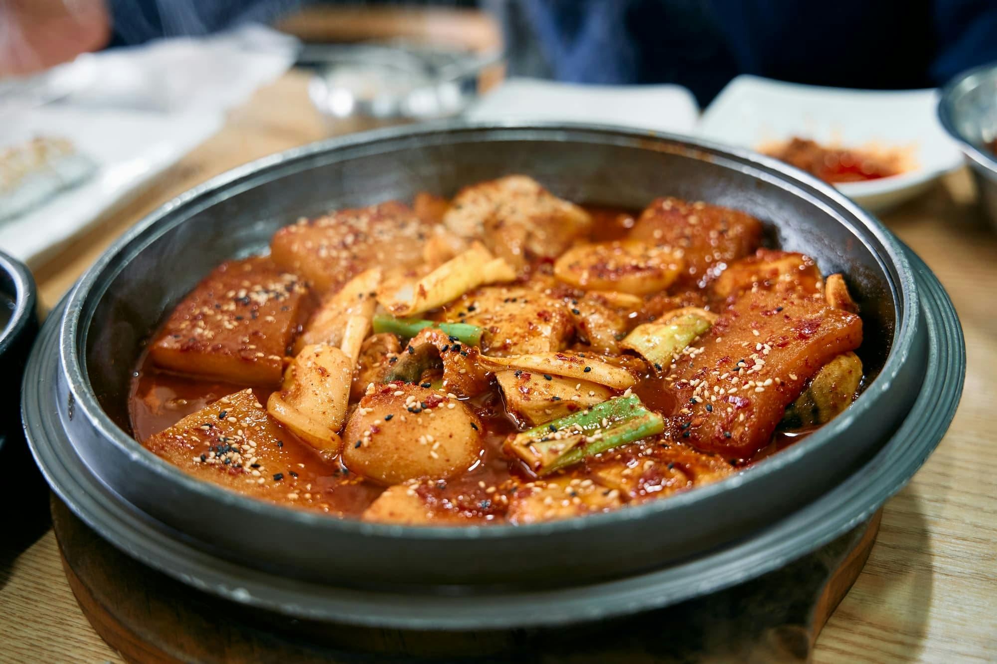 A close-up shot of a dark, metal pot filled with a vibrant orange-red stew, likely Korean in origin. The stew contains chunks of fish or tofu, translucent slices of daikon radish, green scallions, and is generously sprinkled with sesame seeds and red pepper flakes.
