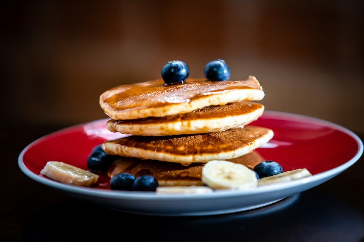 A stack of four golden-brown pancakes sits on a vibrant red plate with a white rim. The top pancake is adorned with two fresh blueberries, and the stack appears to be drizzled with a light syrup or glaze. Scattered around the base of the pancakes on the plate are several more blueberries and slices of ripe banana. The background is softly blurred, suggesting a dark table surface and a warm, out-of-focus backdrop.