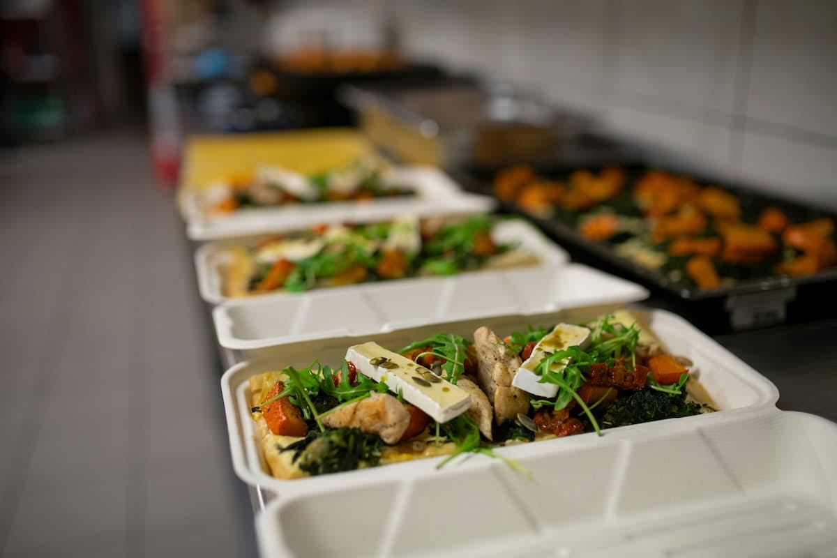 A slightly high-angle, selective focus shot shows a row of white, rectangular takeaway containers filled with prepared meals, likely in a commercial kitchen setting in Rio de Janeiro. The closest container is in sharp focus, revealing a meal consisting of pieces of chicken or fish, vibrant green arugula, roasted orange vegetables (possibly squash or carrots), dark green broccoli florets, and topped with slices of creamy white cheese.
