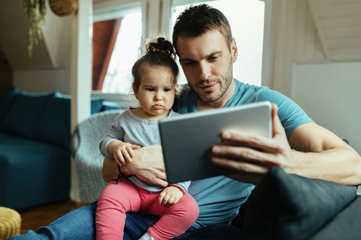 A man, who appears to be a father, sits on a dark couch holding a young child on his lap. They are both looking at a tablet he is holding.