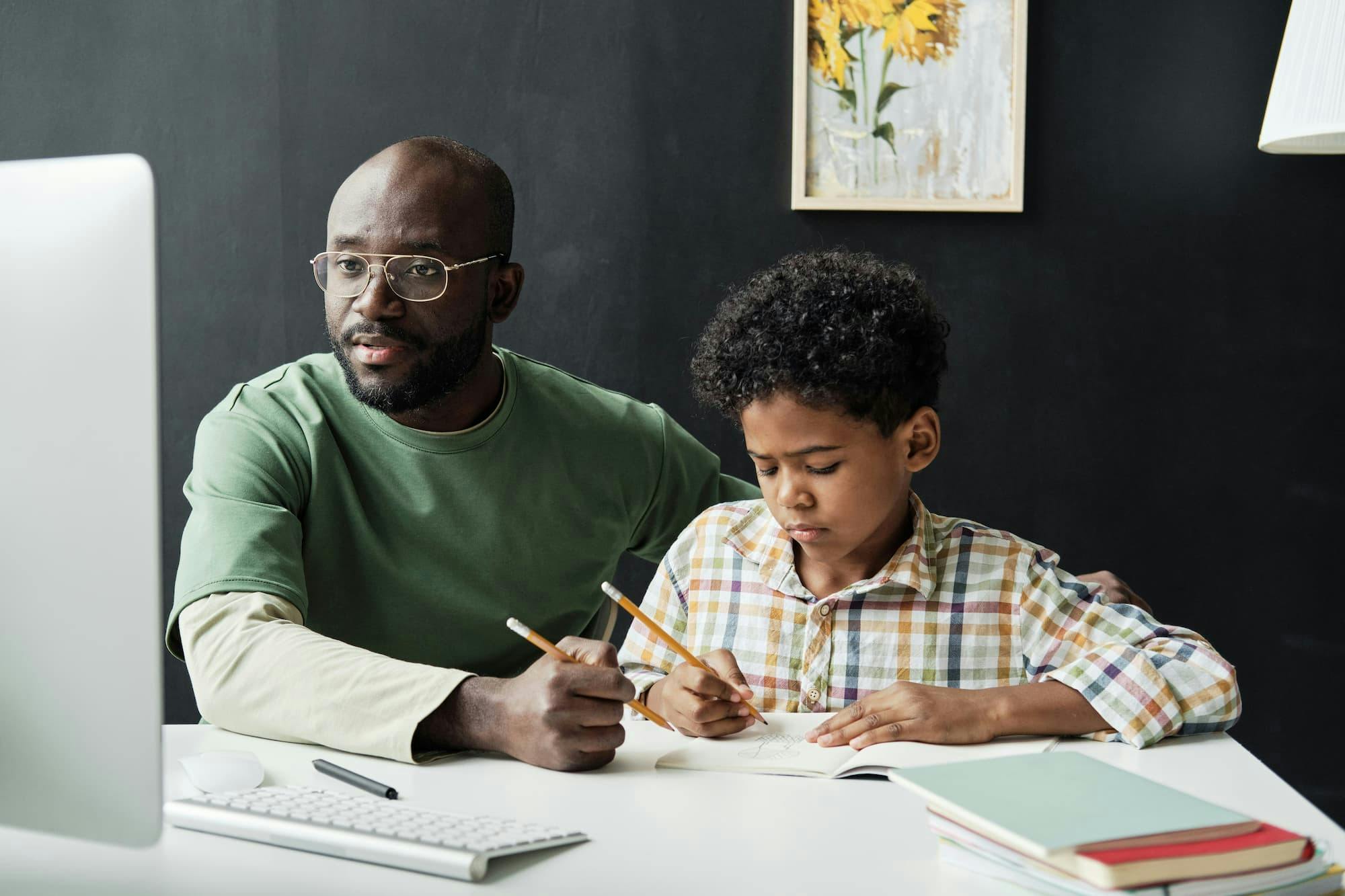 A Black man, likely a father, wearing glasses and a green t-shirt, looks at a computer screen while a young Black boy writes in a notebook at a white desk.