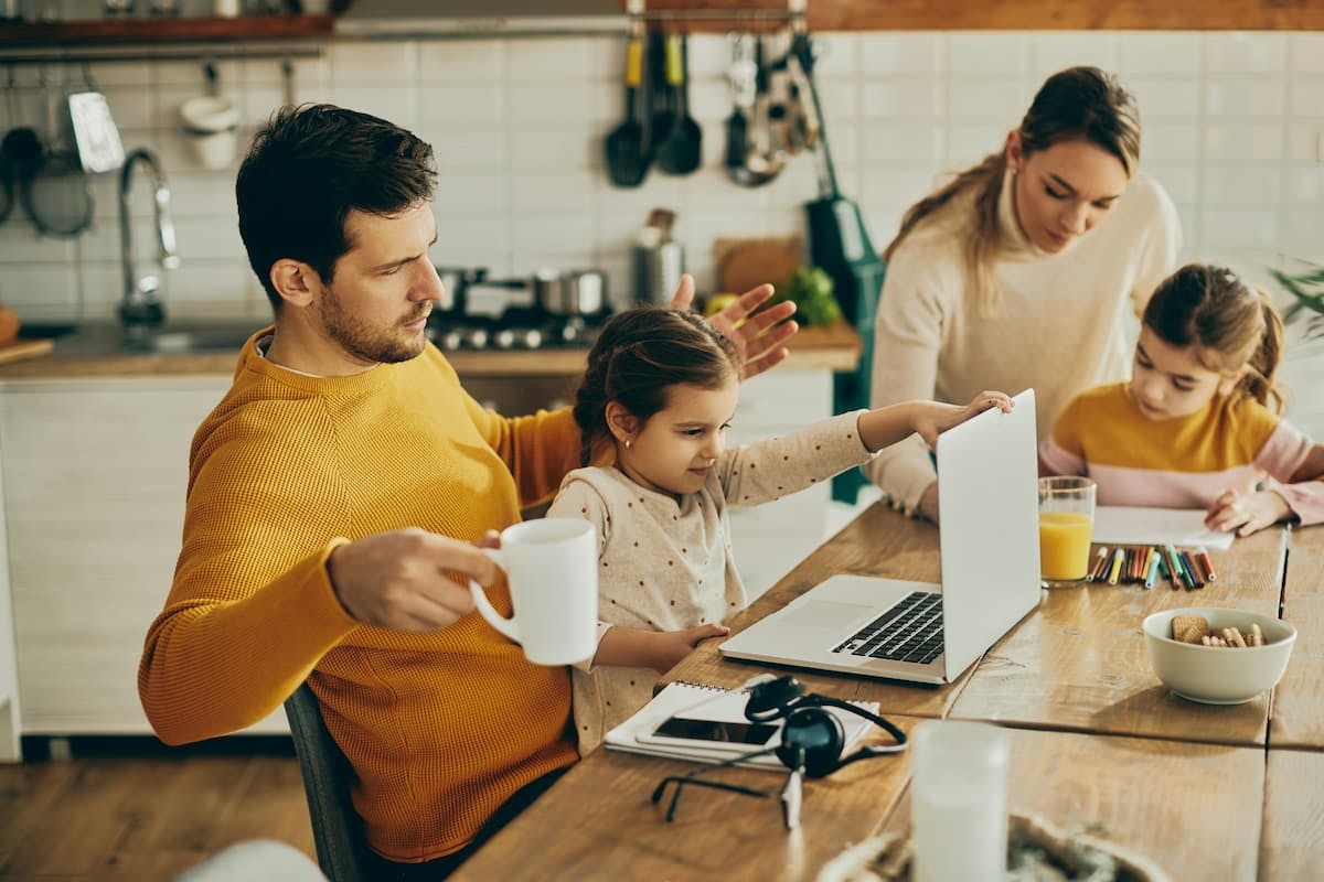 A man, possibly a father, in a yellow sweater sits at a wooden kitchen table holding a white mug and gesturing with his other hand towards a laptop being used by a young girl. Another woman and child are also at the table, the child drawing with crayons.