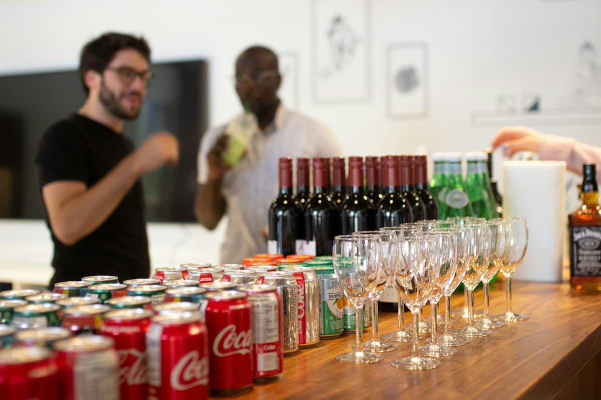 A wooden counter is laden with various beverages, including rows of Coca-Cola cans, several bottles of red wine, green bottles (possibly sparkling water or soda), numerous wine glasses, and a bottle of Jack Daniel's. In the blurred background, two individuals appear to be conversing in an office setting, suggesting a Father's Day office party.