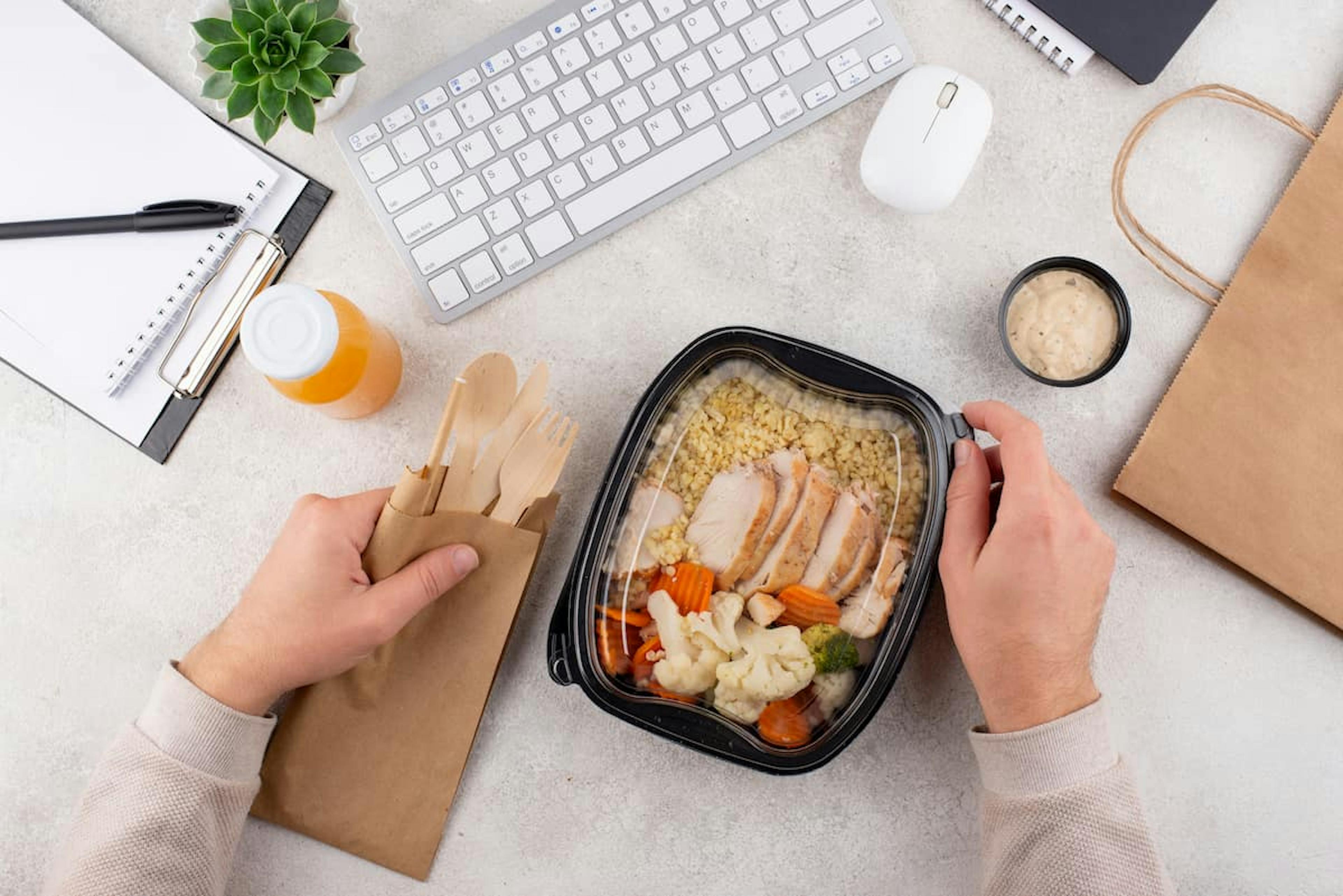 A person enjoying a meal at a desk with a laptop and a bag of food, illustrating office meal delivery options.