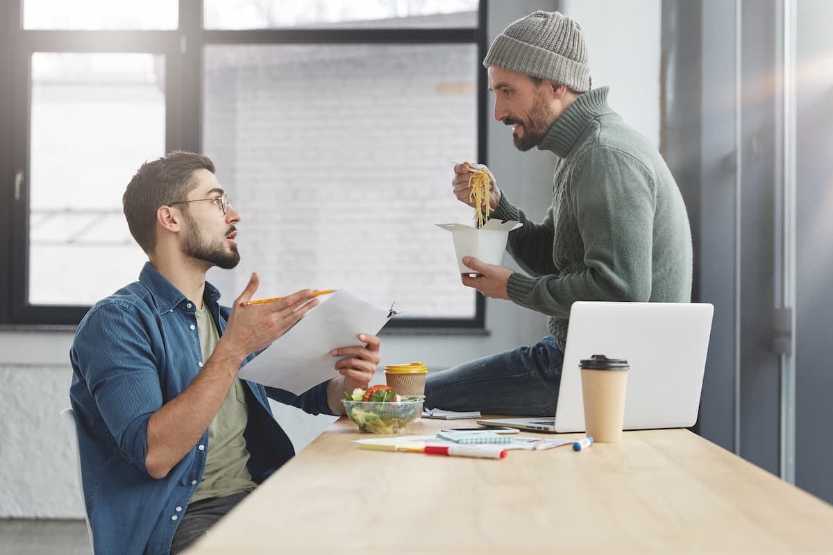 Two men enjoying a meal together at a table, discussing options for an office meal delivery service.