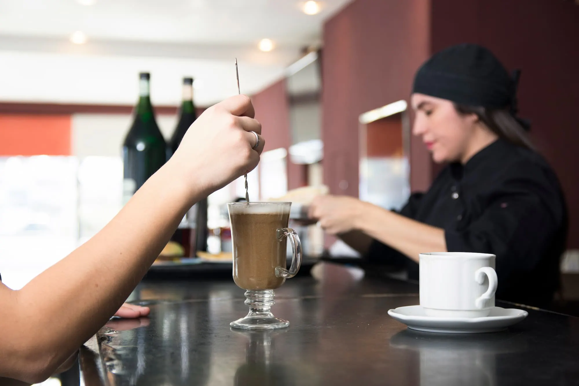 Person stirring coffee at a counter with a barista in the background, showing in-house cafeteria vs. meal delivery experience.