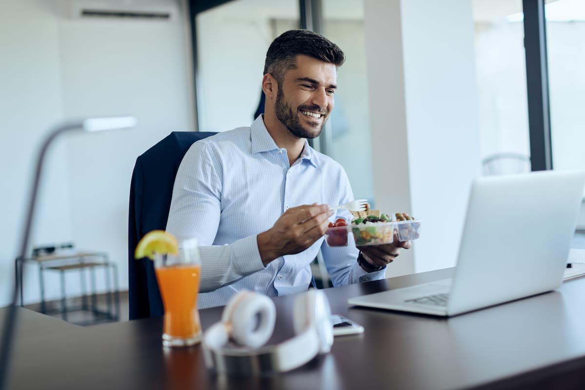 Smiling man eating salad and tomatoes at his desk, showing one of the most popular office lunch orders.