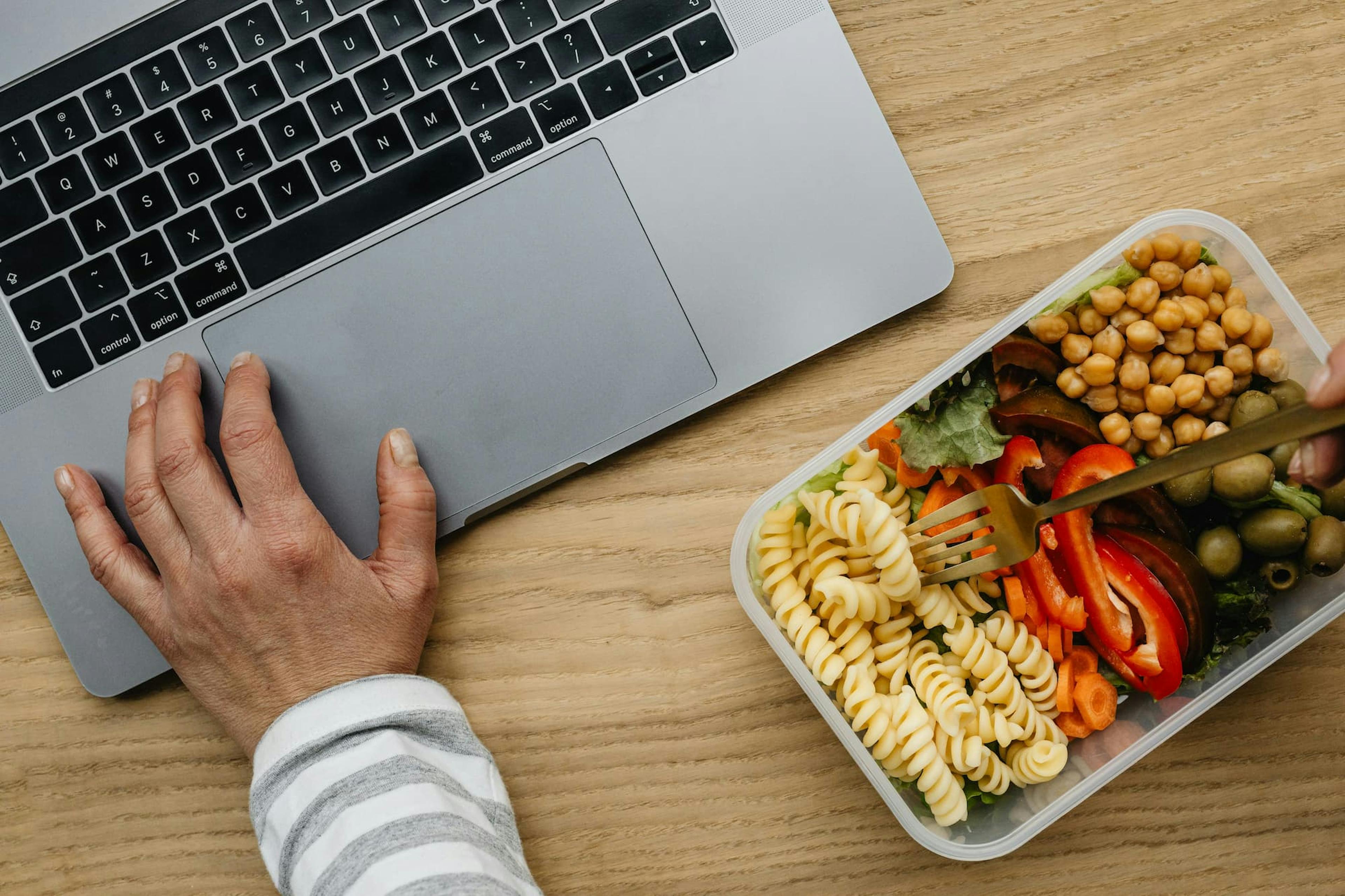 Person eating homemade lunch while working on laptop, illustrating how to save money on office lunches with meal prepping.