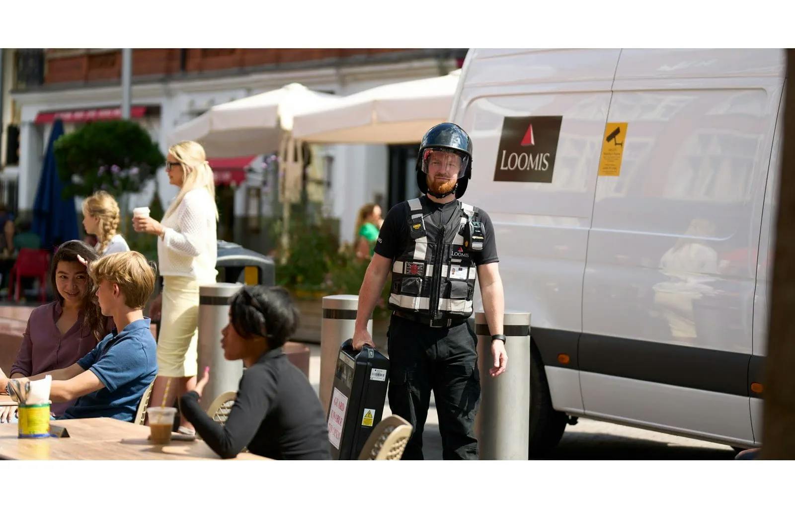 Loomis-väktare i skyddsutrustning bär värdetransportväska intill ett Loomis-fordon i stadsmiljö. Loomis är ett av världens ledande bolag inom kontanthantering och värdetransport.