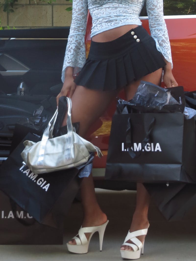 A model wearing a short black skirt, light blue lace top, and white heels is holding multiple black shopping bags labeled "I.AM.GIA" and a silver handbag. They are standing in front of the open door of a red car.