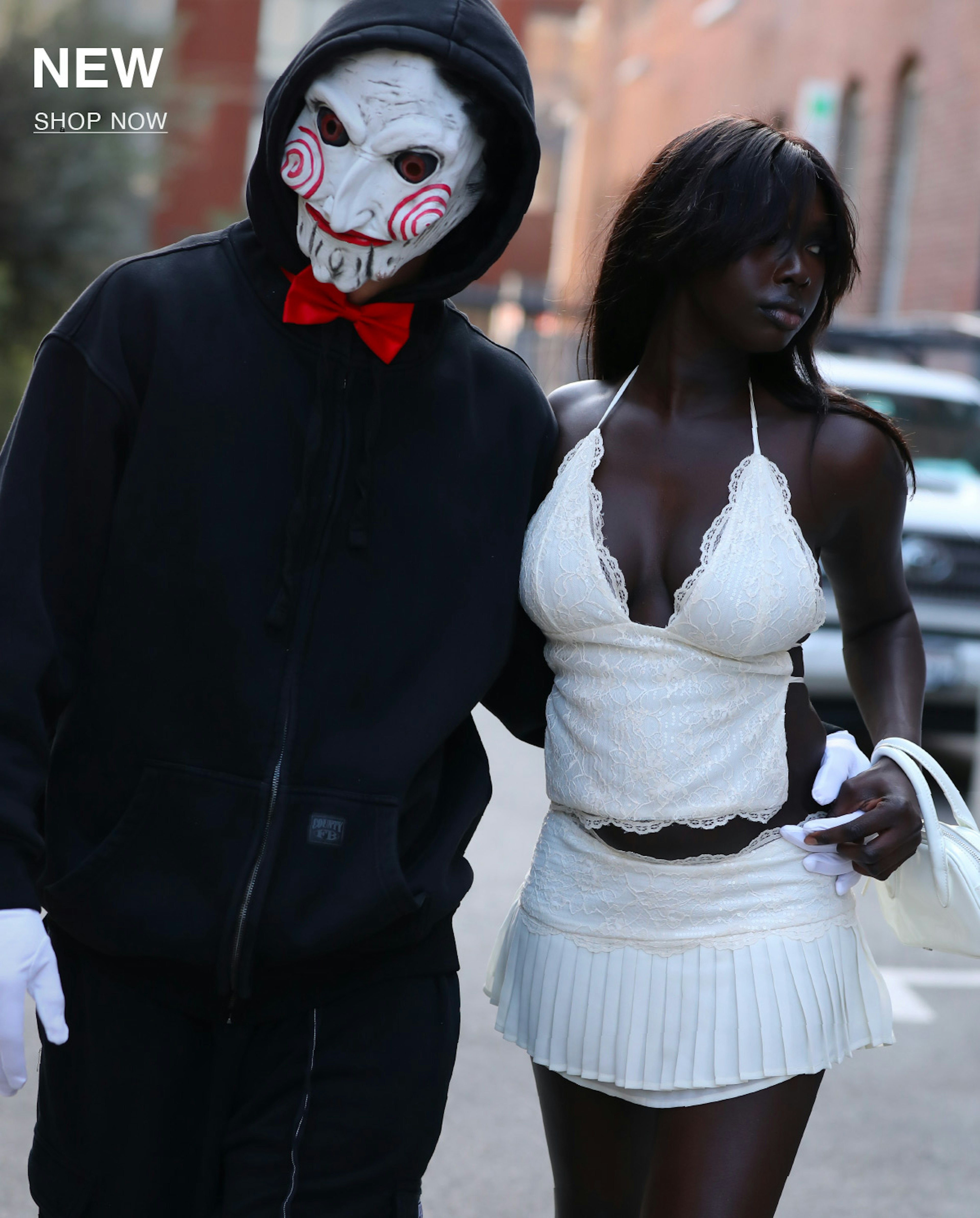 A person in a black hoodie and white Jigsaw mask stands next to a woman wearing a white lace halter top and pleated mini skirt as they walk outside on a city street. Text in the corner reads "NEW SHOP NOW.