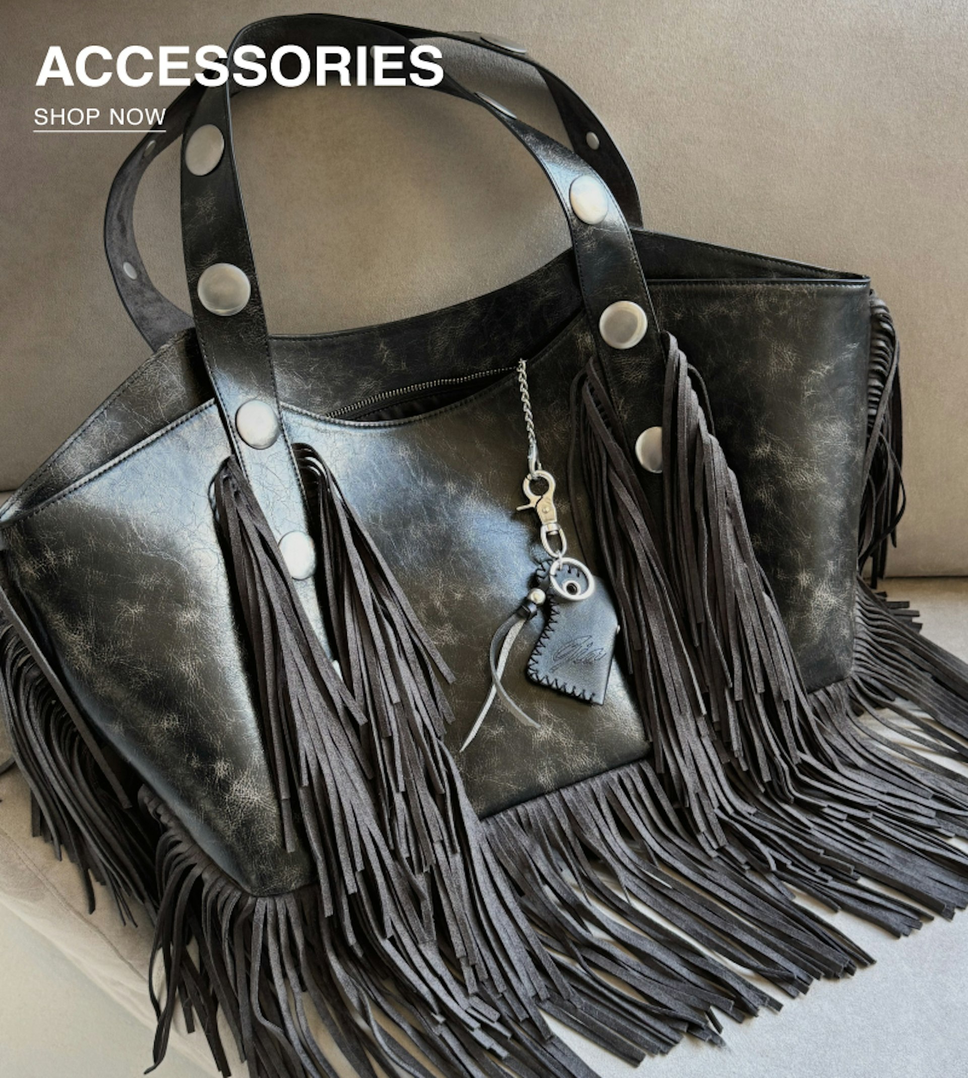 A black leather fringe handbag with silver studs and a keychain charm rests on a beige surface. Text in the corner reads “ACCESSORIES SHOP NOW.”.