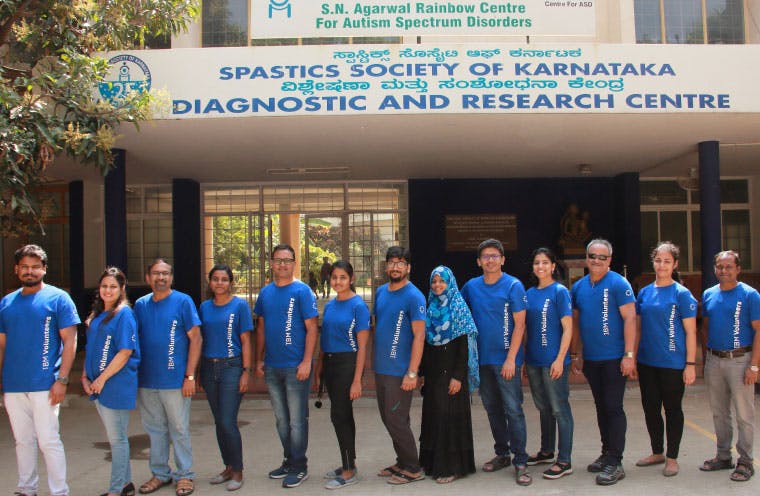 A group of people standing in a row under at the entrance to a building with two signs over the door. One reads, S.N. Agarwal Rainbow Centre for Autism Spectrum Disorders. The other sign reads, "Spastics Society of Karnataka, Diagnostic and Research Centre."