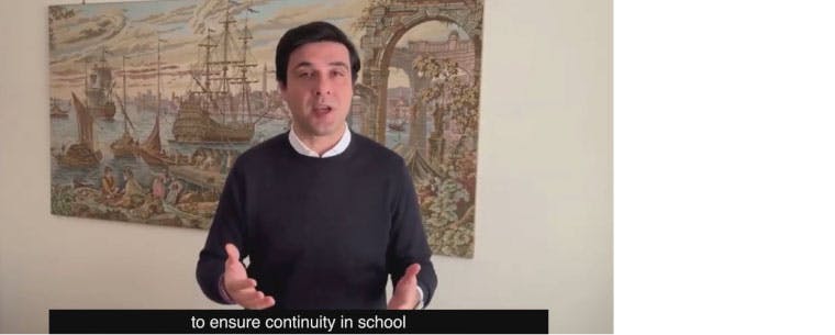 A screenshot taken from a video of a man in front of painting of an Italian river scene. The subtitle at the bottom of the screen shot reads, "to ensure continuity in school"