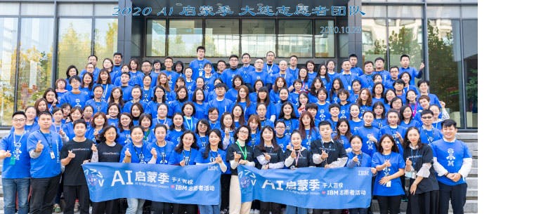 A very large group of people arranged in rows on steps. Most are wearing blue t-shirt with the pictogram of an eye, a bee and the letter M. The front row holds two banners reading IBM volunteers and AI Enlightenment Season