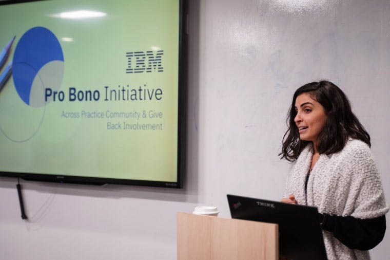 A woman speaks from behind a lecture, the screen beside her reads "IBM Pro Bono Initiative, Across Practice Community & Give Back Involvement
