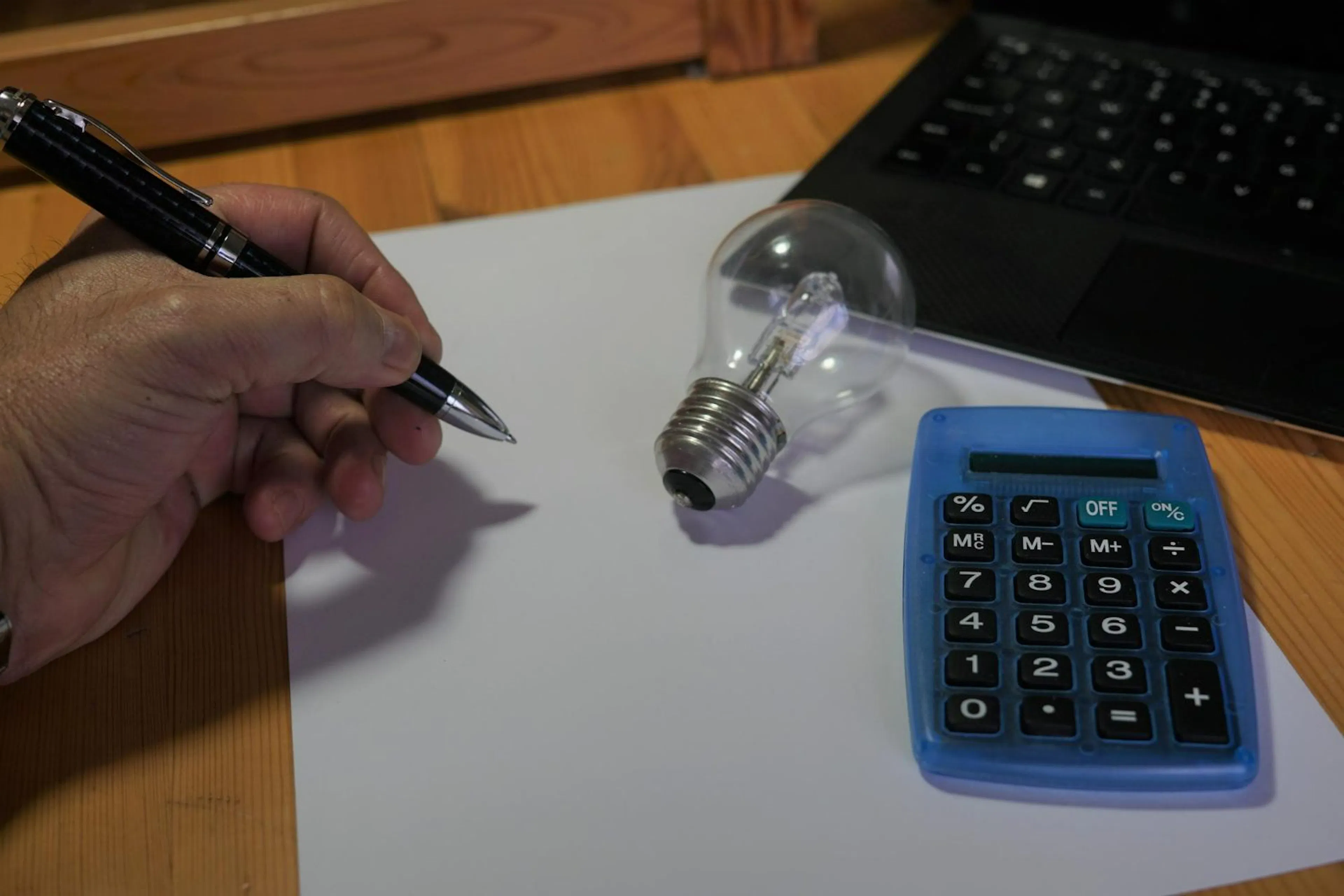 Hand holding a pen beside a calculator and a light bulb on a desk
