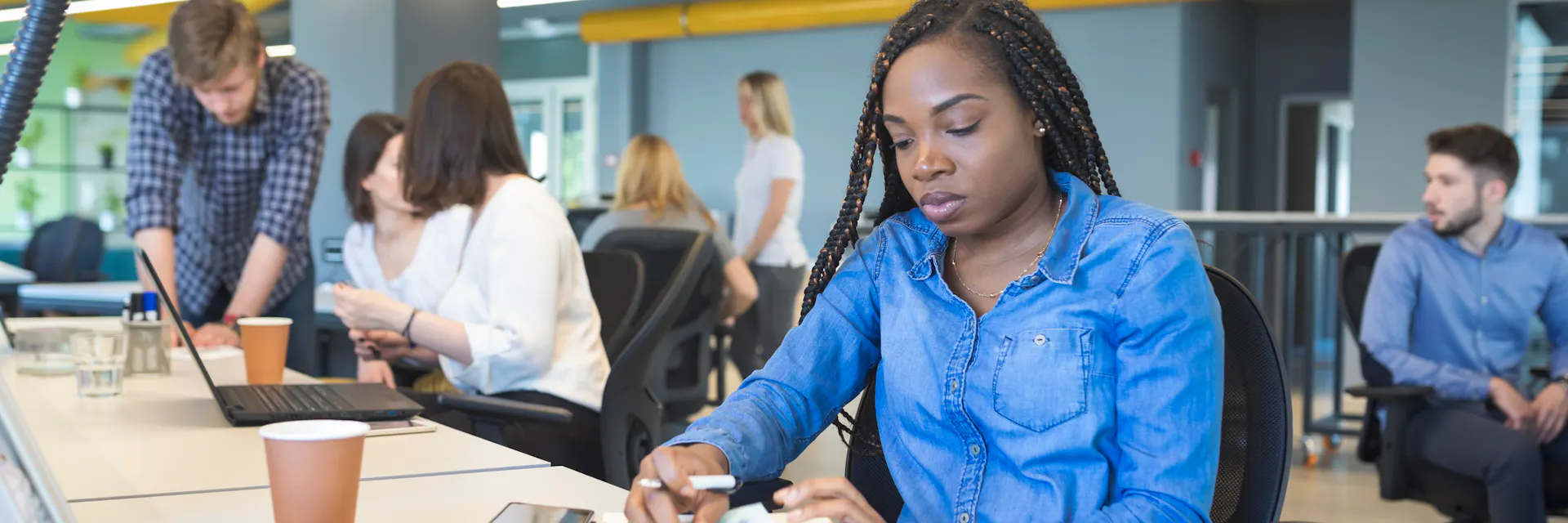 A young black woman sits at a desk in an open plan office area surrounded by young white colleagues who are interacting among themselves, but not with her.