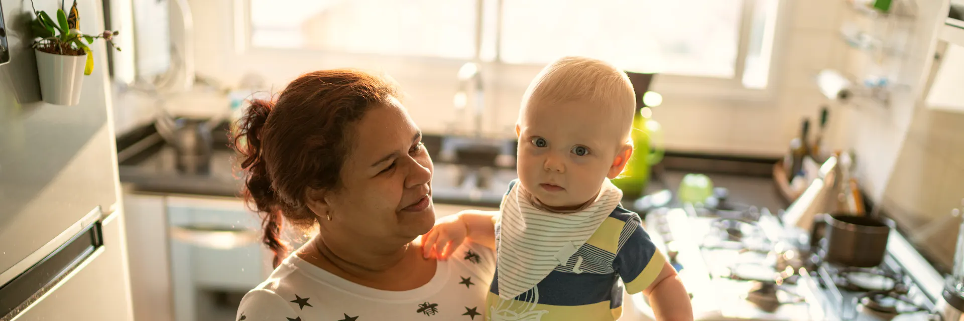 Nanny in Brazil holds baby in her arms. They are in the kitchen.