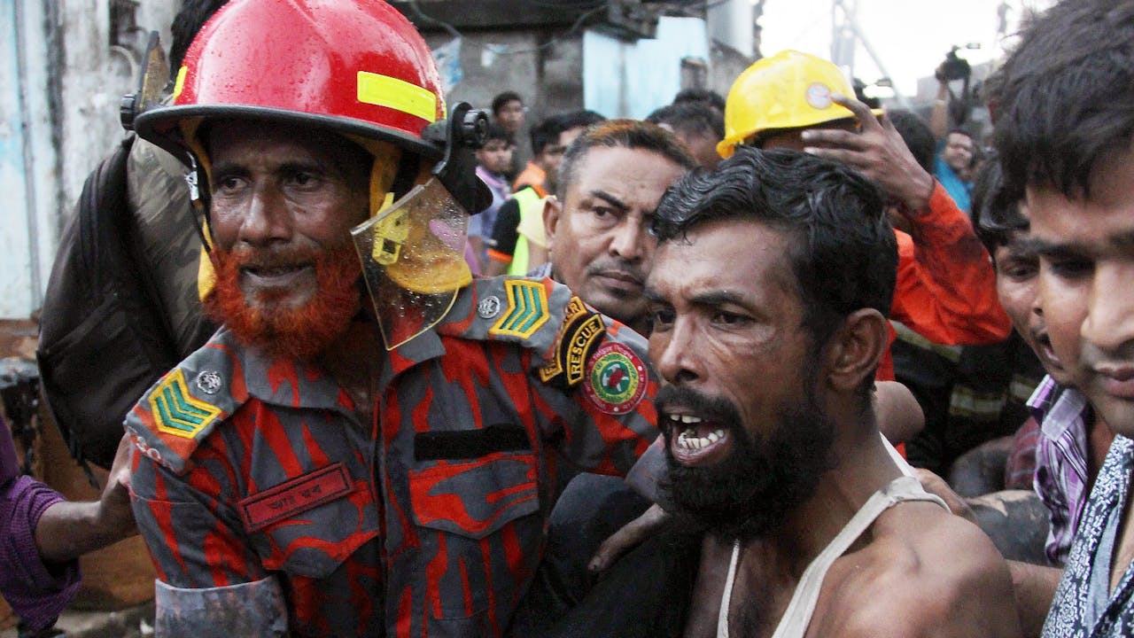 Mohamed Abdur Rob in firefighter uniform and red hard hat carries an injured victim after a fire incident in Tongi, Dhaka. A crowd of agitate people surround him.