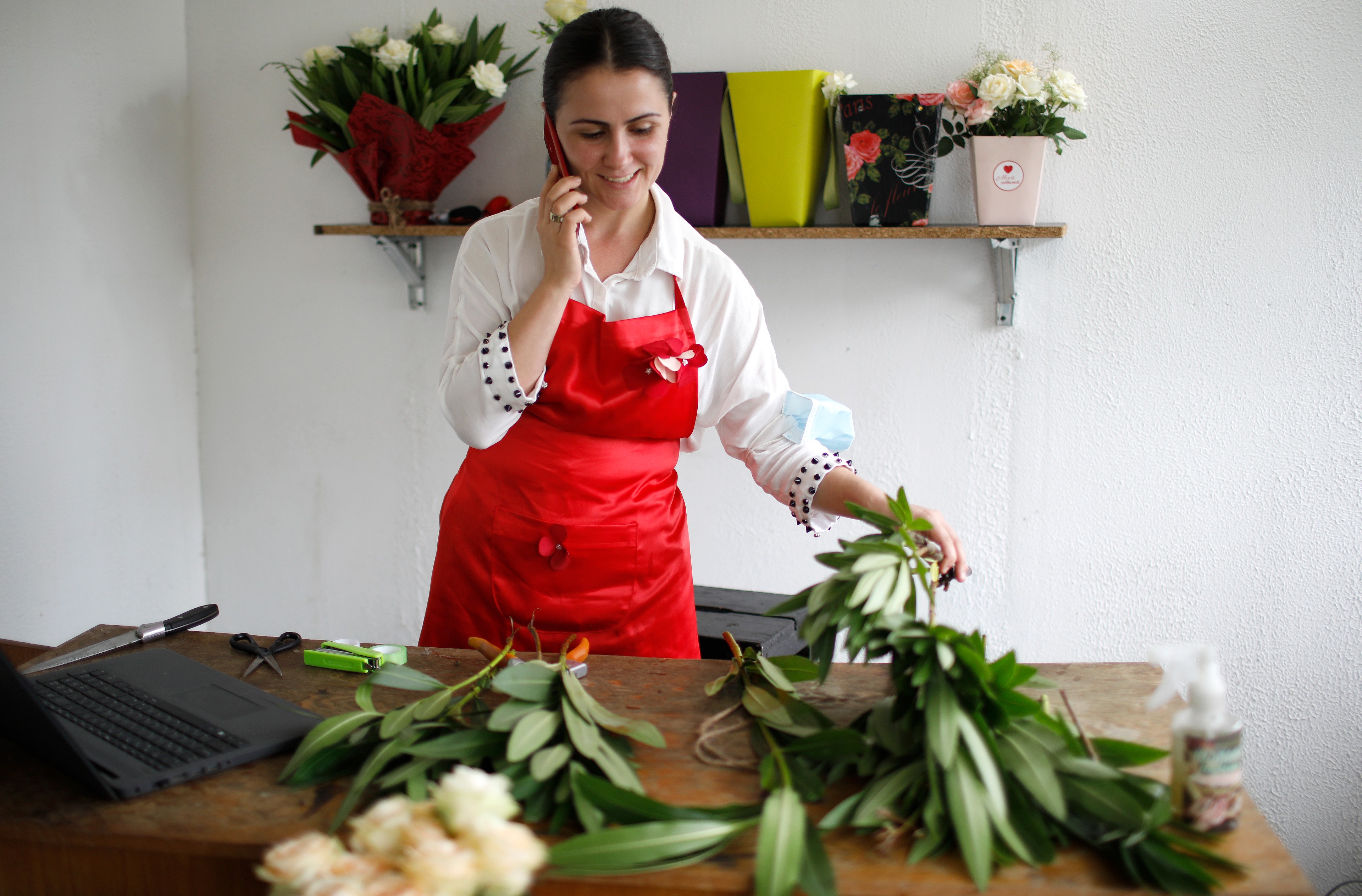 Mariam Kobalia stands in her flower shop, holds her phone in one hand and a flower in the other.   In the foreground on the table there are cut flowers and a laptop.