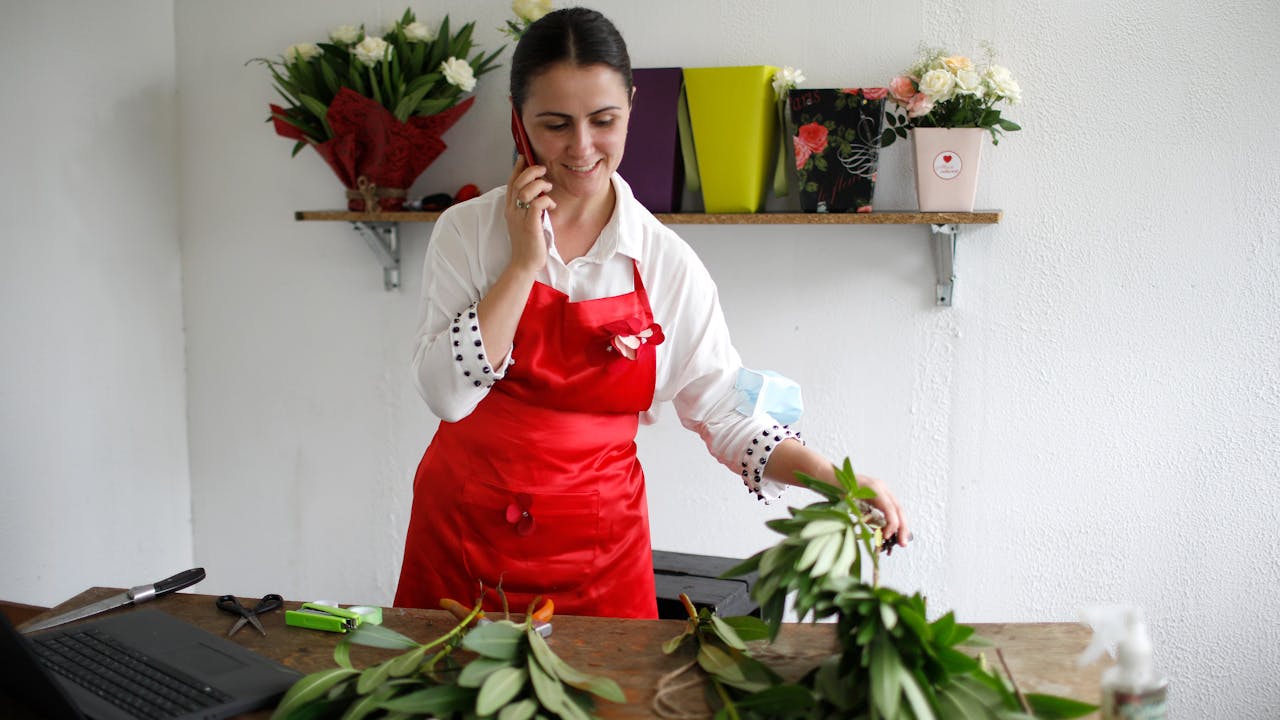 Mariam Kobalia stands in her flower shop, holds her phone in one hand and a flower in the other.   In the foreground on the table there are cut flowers and a laptop.