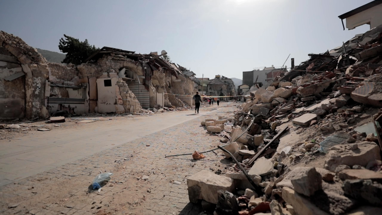 A man stands in the middle of a street in Hatay province in Türkiye a few days after the earthquakes have taken place. Either side of the street are collapsed buildings and rubble.