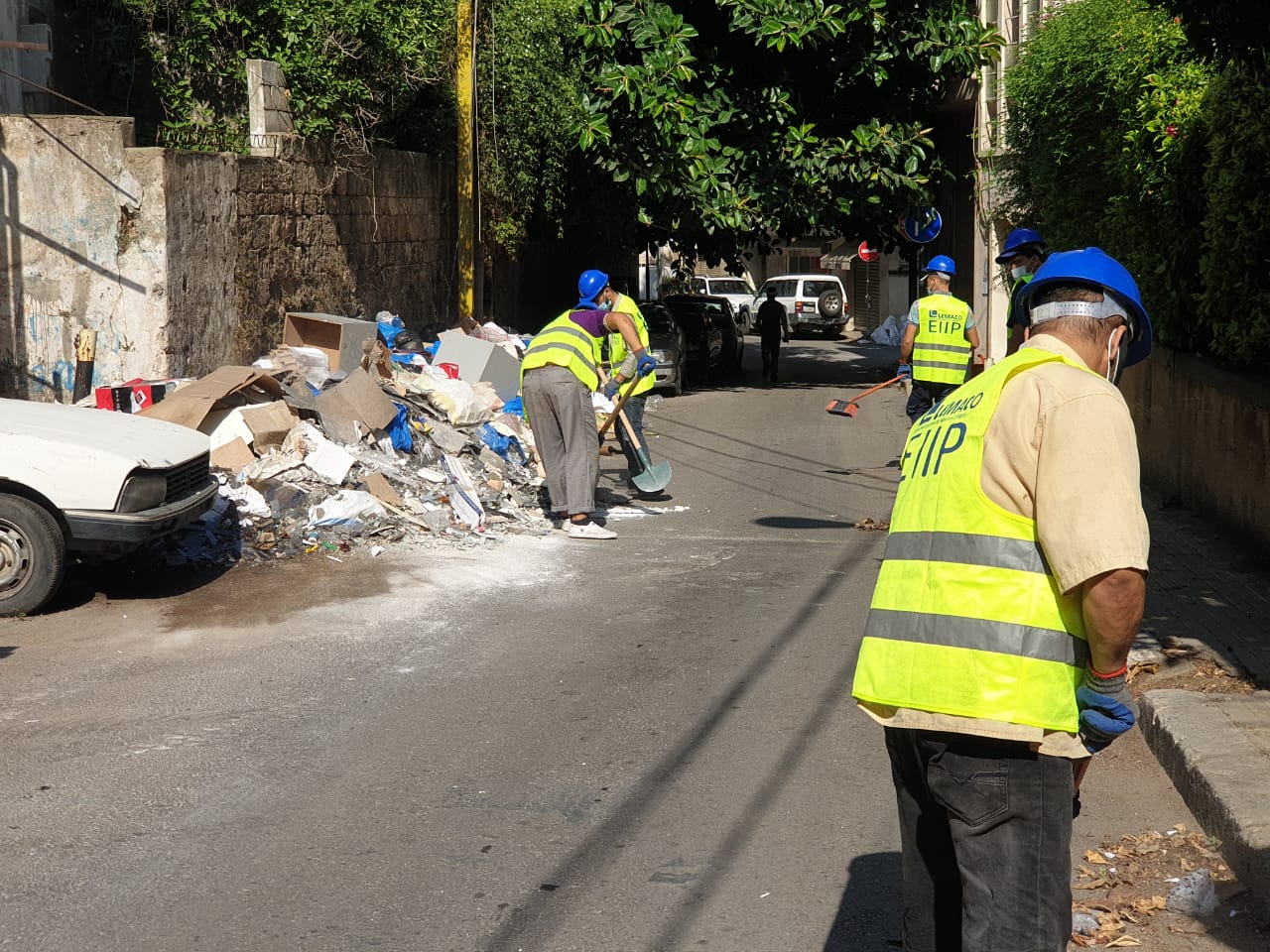 Workers clear rubble on Beirut street and wear blue hard hats and fluorescent jerseys with EIIP written on them.